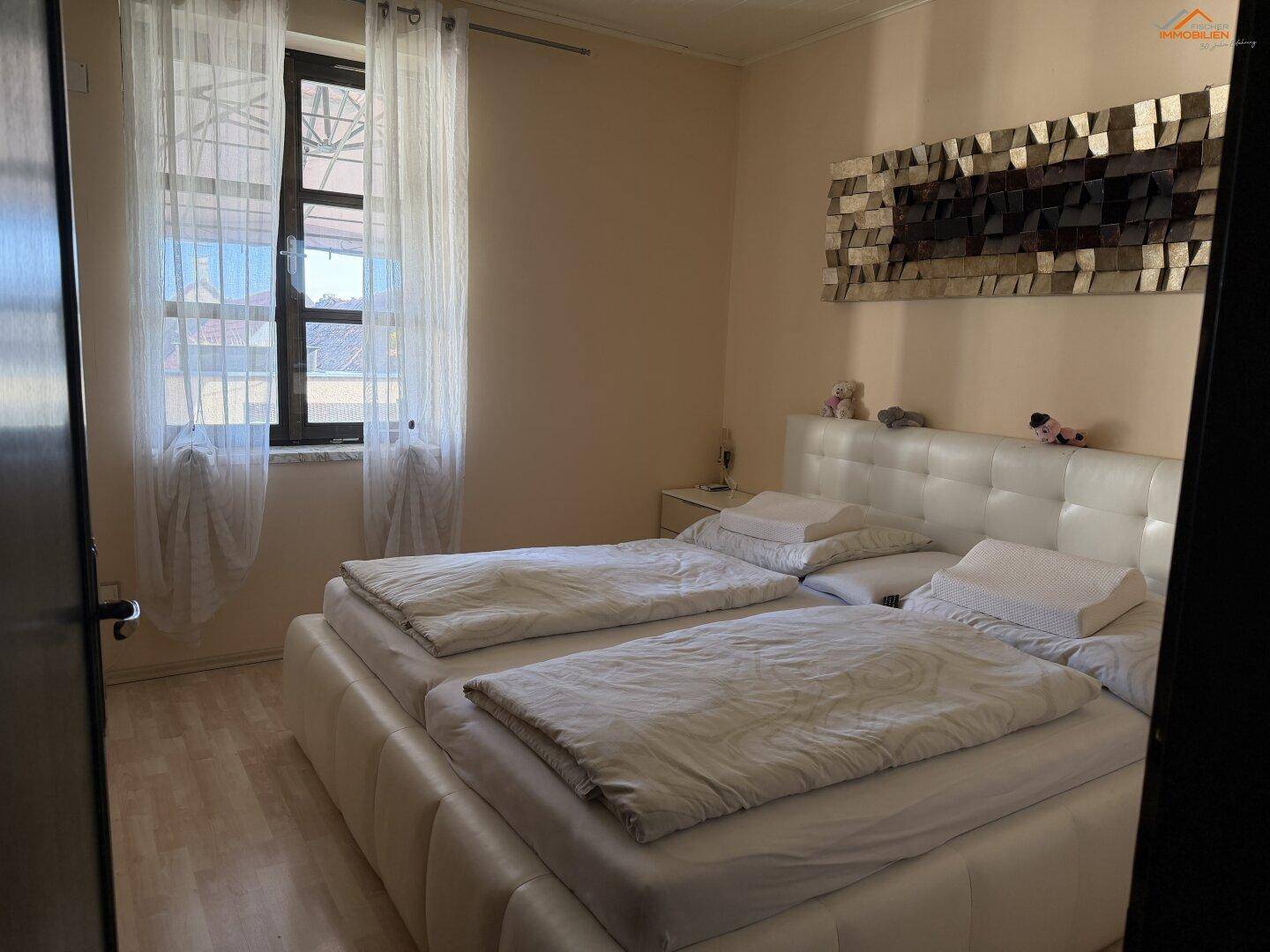 Schlafzimmer mit weißem Polsterbett, hellem Holzboden und Fenster mit Vorhängen.
