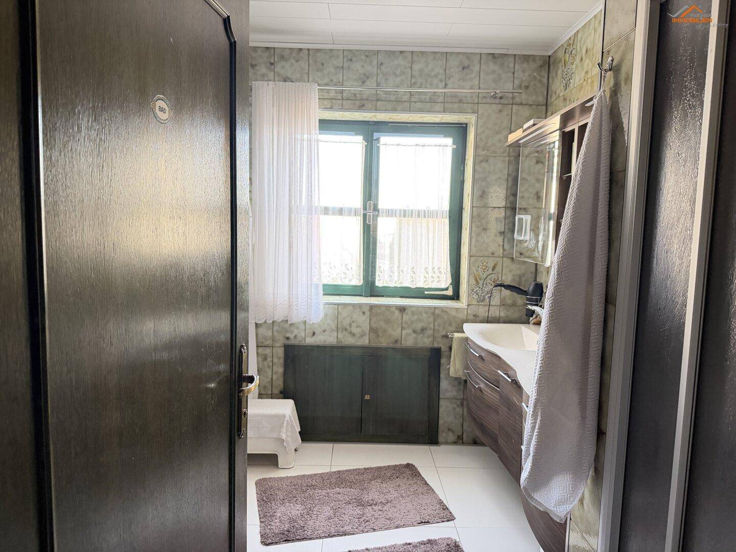 Badezimmer mit Waschbecken, Spiegelschrank und Fenster, gefliest in Grün- und Beigetönen.