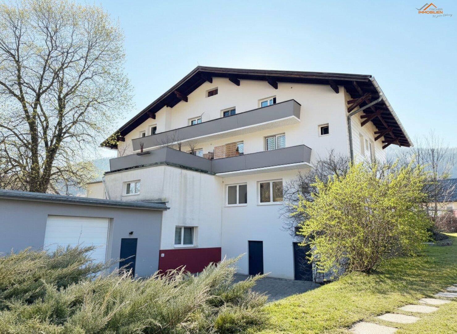 Rückansicht des Gebäudes mit Balkonen, angrenzender Garage und Gartenbereich vor bewaldeten Hügeln.
