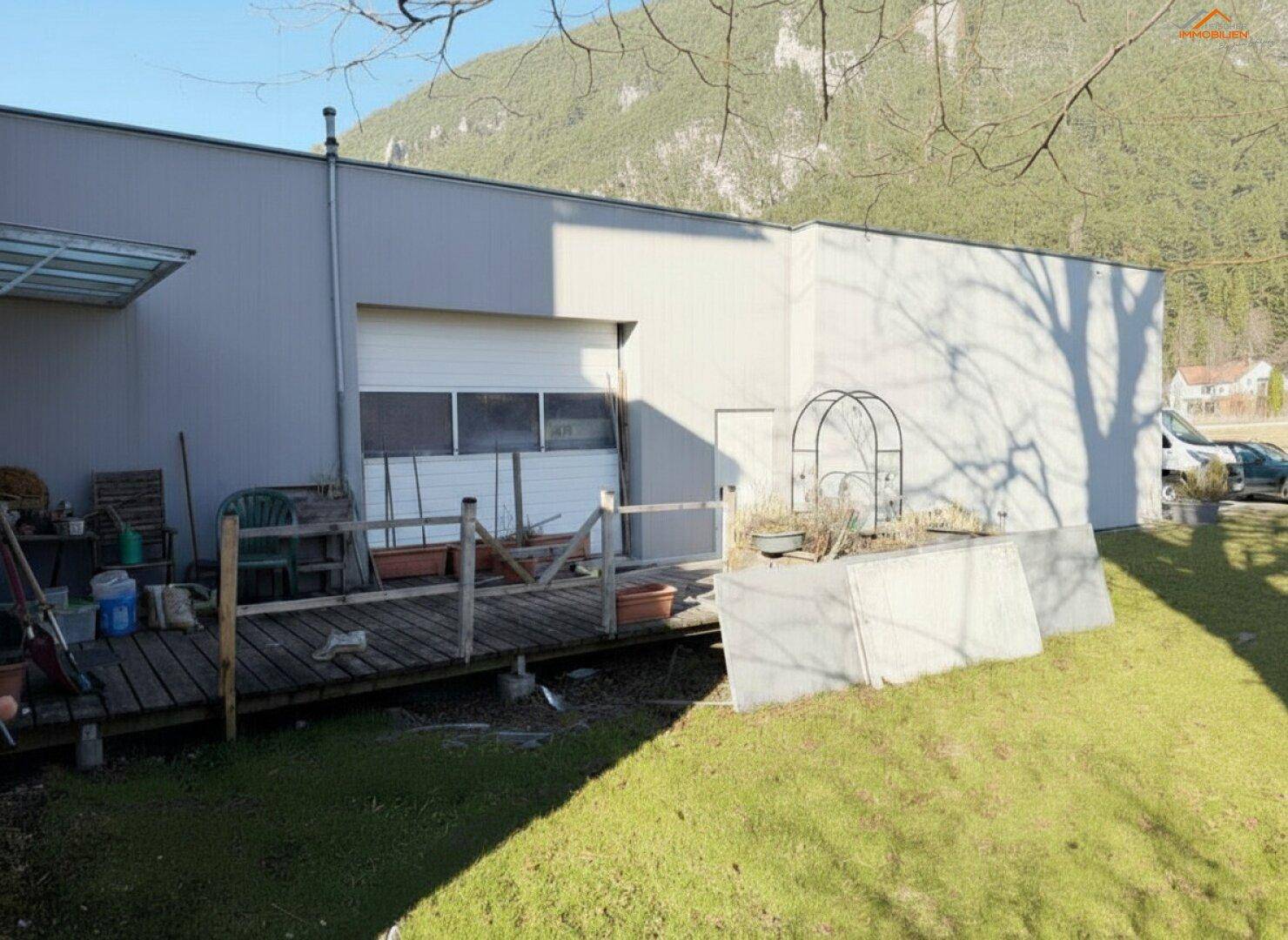 Industriell anmutendes Nebengebäude mit Garagentor, Holzterrasse und Grünfläche vor einem Bergpanorama.
