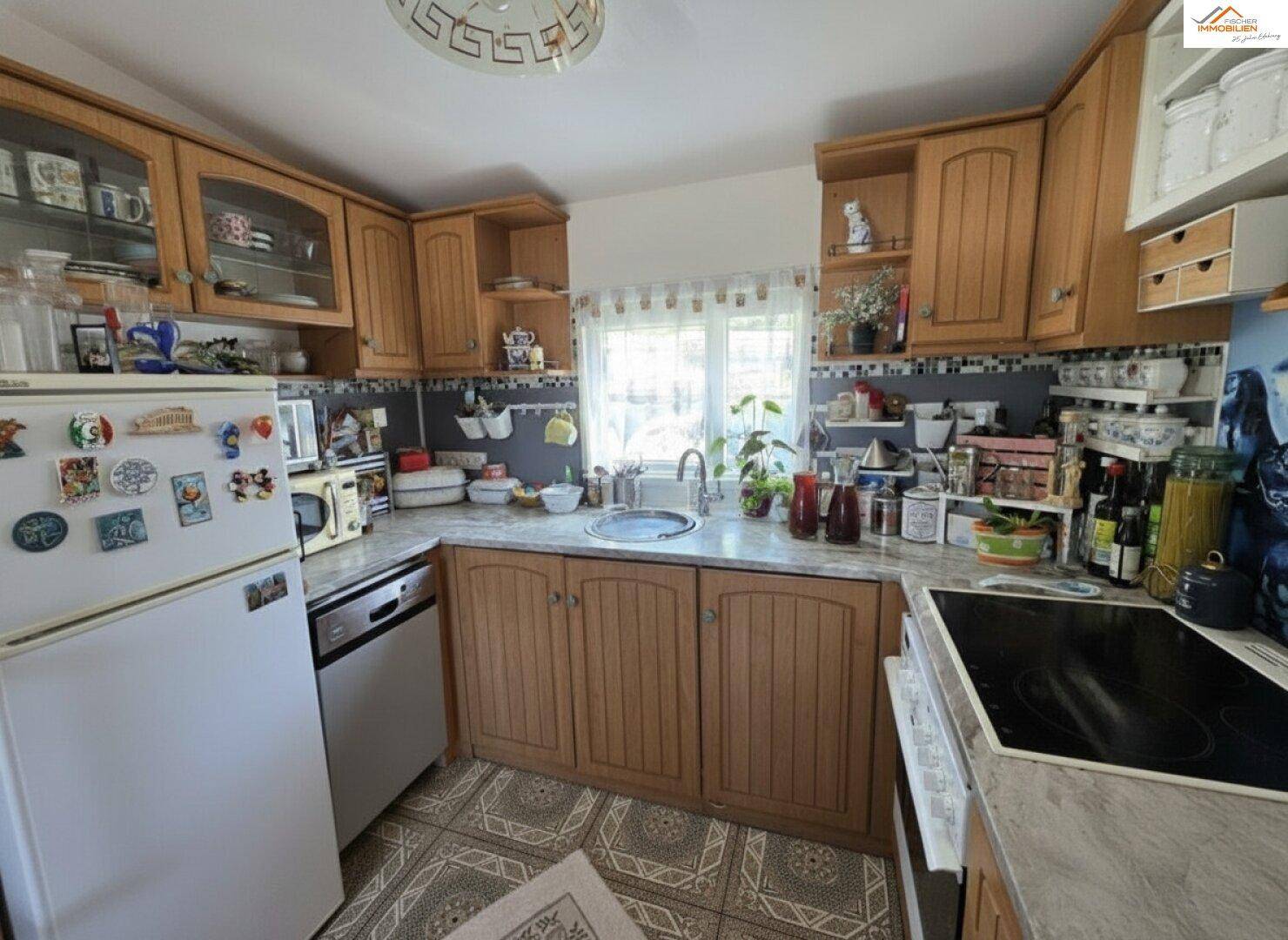 Funktionale Küche mit Holzschränken, Fliesenboden und Fenster über der Spüle, ausgestattet mit grundlegenden Geräten.
