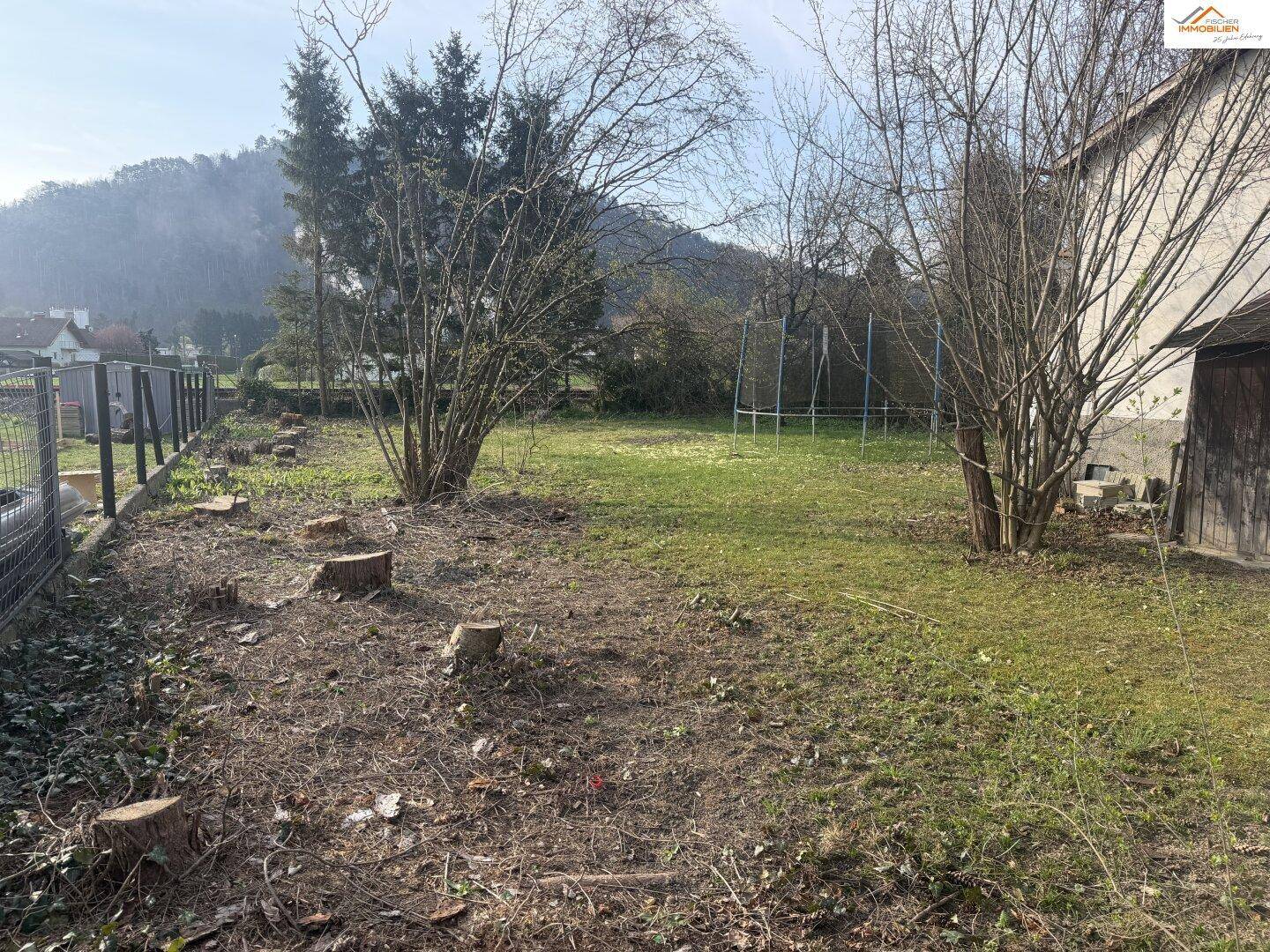 Gartenfläche mit Baumstümpfen und einem Trampolinrahmen, umgeben von Bäumen und Hügeln.