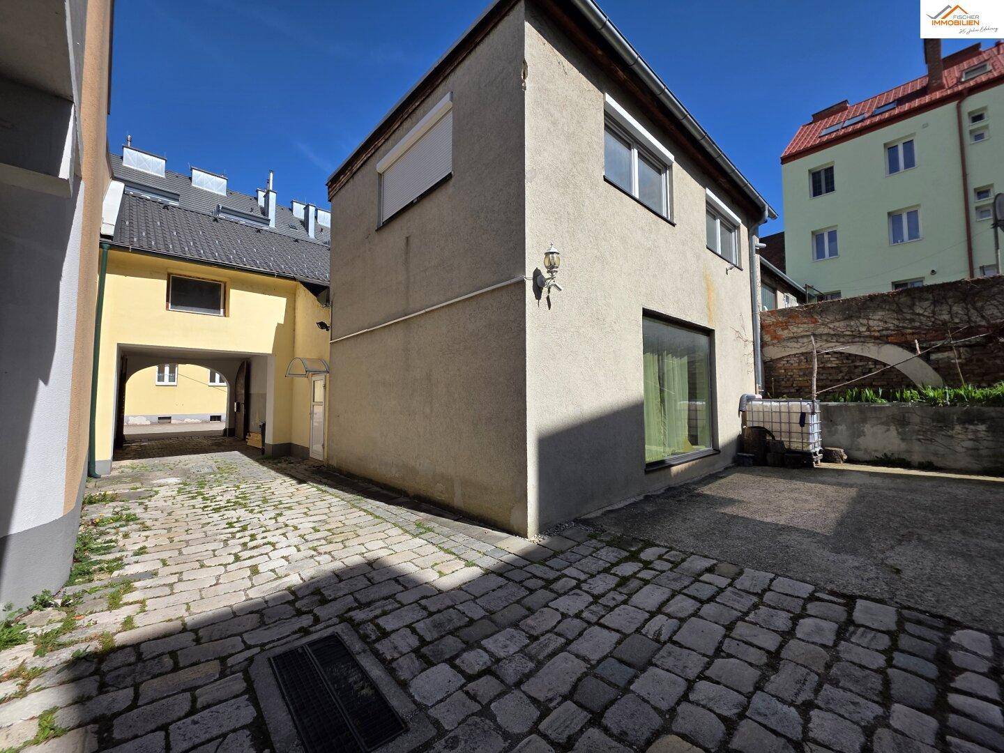 Hinterhofbereich mit Kopfsteinpflaster und einem Gebäude mit großen Fenstern im Erdgeschoss.