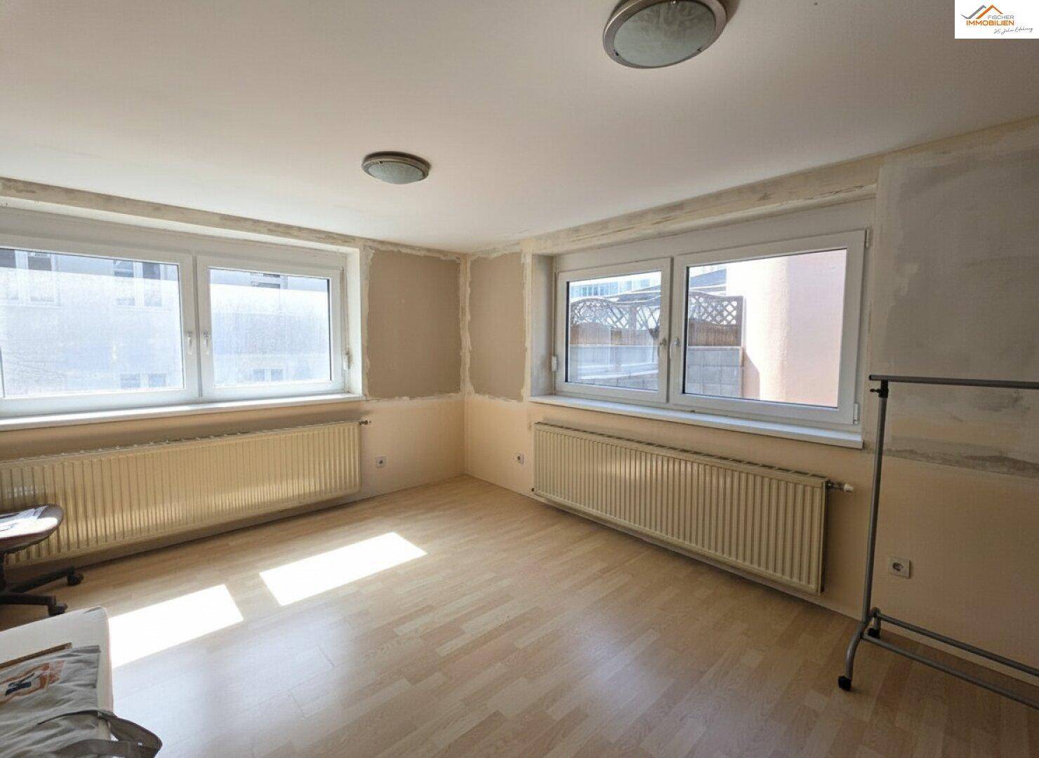 Heller Raum mit Laminatboden und großen Fenstern, Wände in Vorbereitung für Renovierung.