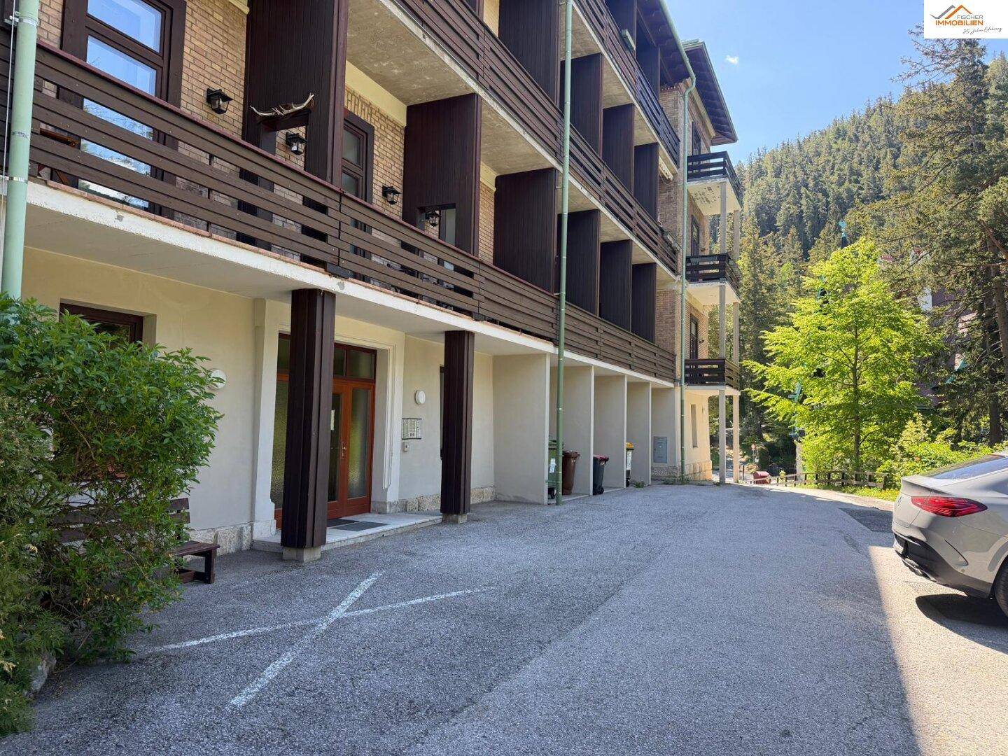 Eingangsbereich des Gebäudes mit Holztür, Säulen und einem geparkten Auto auf dem Vorplatz.