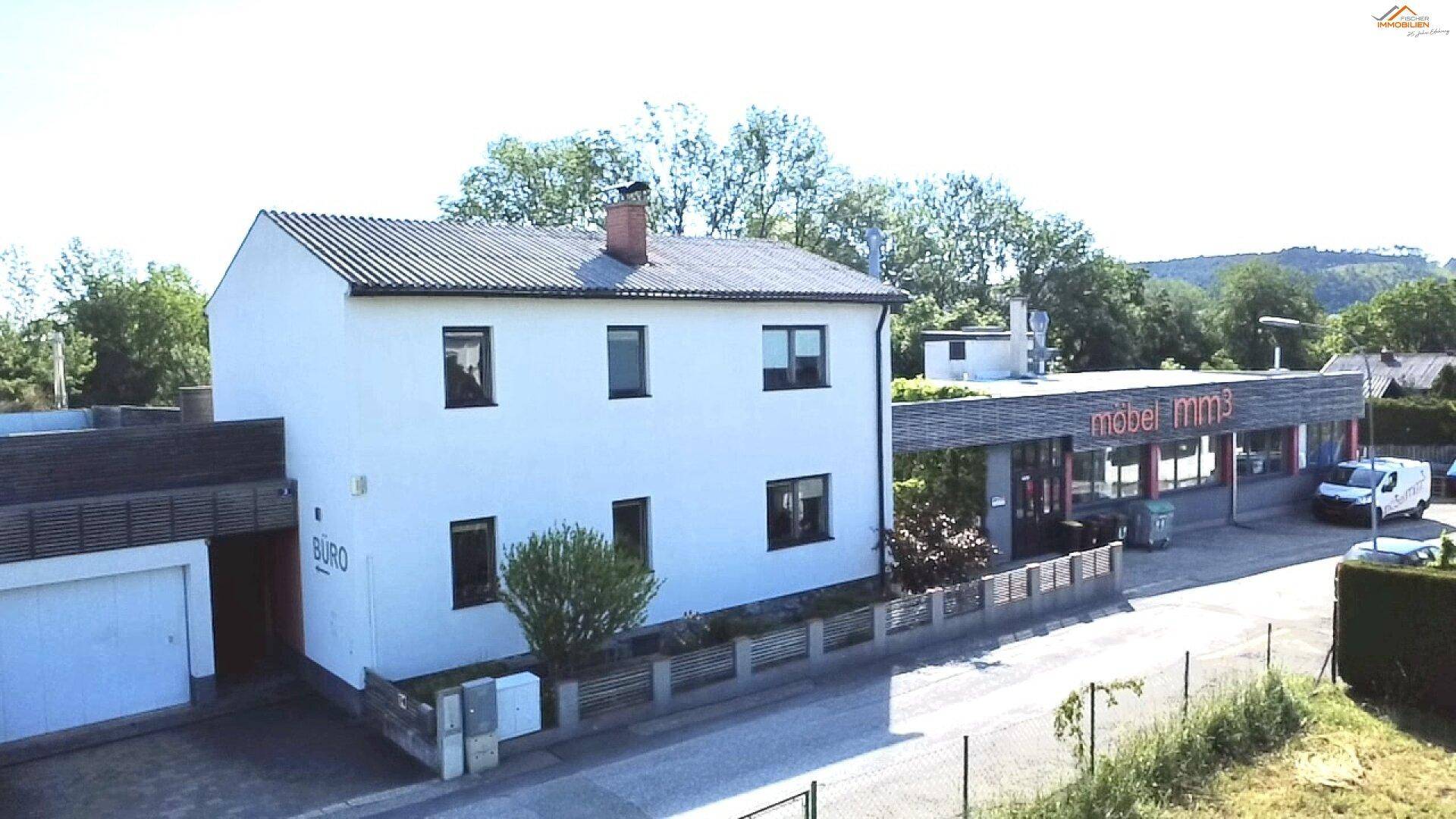 Luftaufnahme des weißen Hauses mit "BÜRO"-Schild an der Garage, Holzterrasse und dem angrenzenden Gewerbegebäude bei Sonnenschein.
