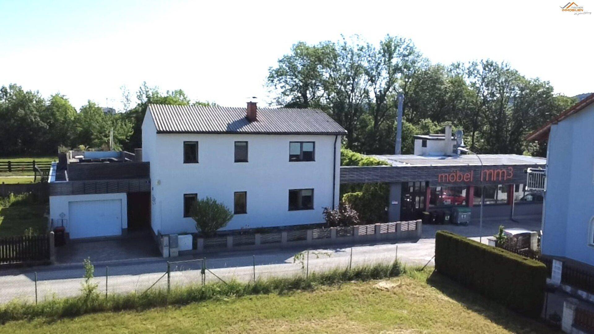 Luftaufnahme des weißen Hauses und des Gewerbegebäudes, mit einer Grünfläche im Vordergrund und einem blauen Haus im Hintergrund.