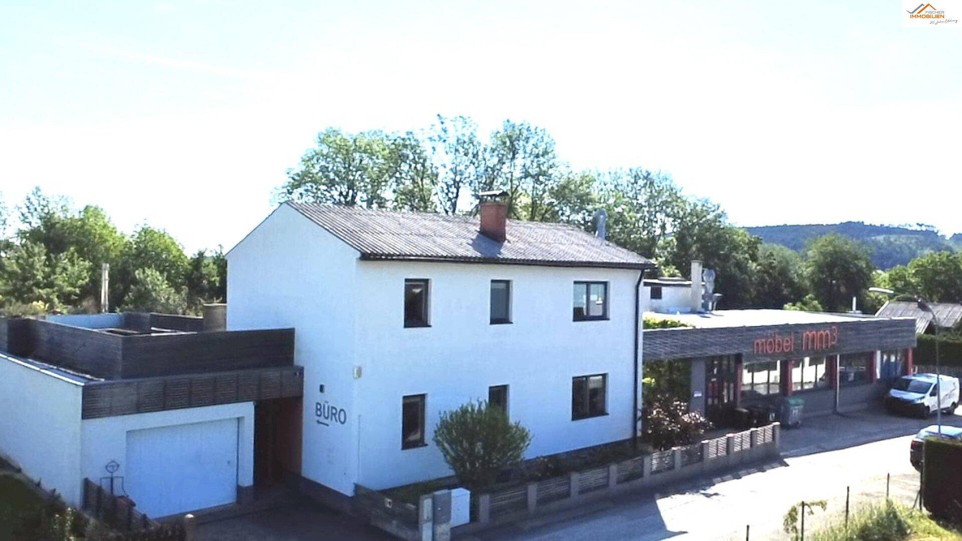 Luftaufnahme des weißen Hauses mit "BÜRO"-Schild an der Garage, Holzterrasse und dem angrenzenden Gewerbegebäude.