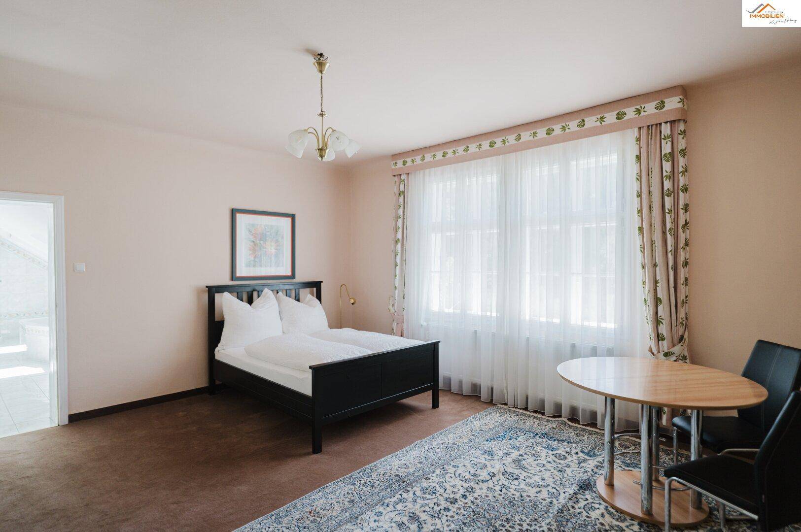 Schlafzimmer mit Doppelbett, kleinem Esstisch, Teppichboden und großen Fenstern mit Vorhängen.