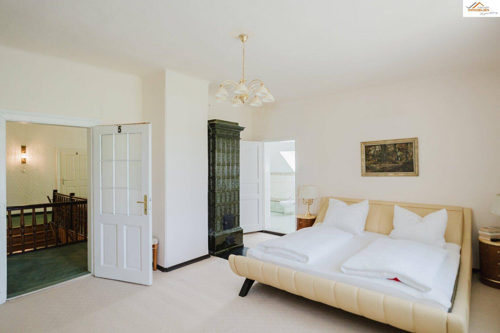 Helles Schlafzimmer mit Doppelbett, traditionellem Kachelofen und Zugang zu einem Treppenhaus.