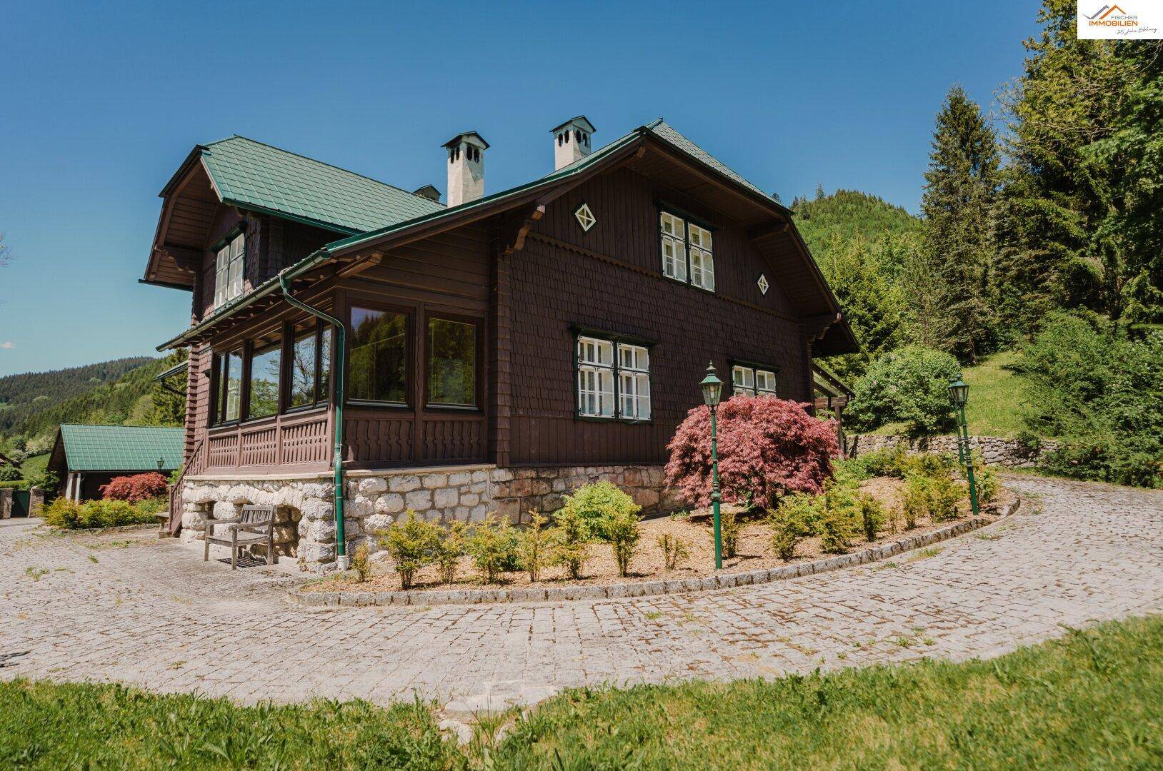 Traditionelles Holzhaus mit grünem Dach, weißen Fenstern und gepflasterter Auffahrt in sonniger Umgebung.
