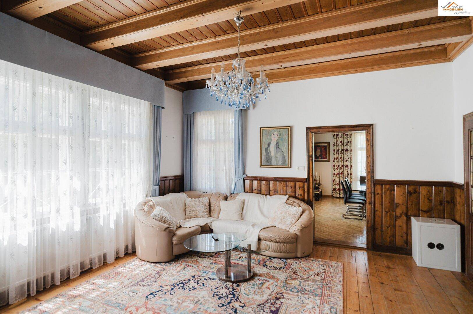 Gemütliches Wohnzimmer mit großem Ecksofa, Holzbalkendecke und einem eleganten blauen Kronleuchter.