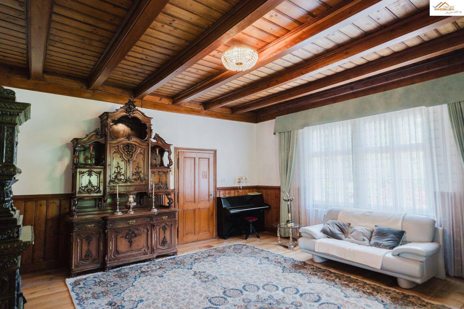 Geräumiges Wohnzimmer mit Holzbalkendecke, weißem Sofa, antikem Holzschrank und einem Klavier.