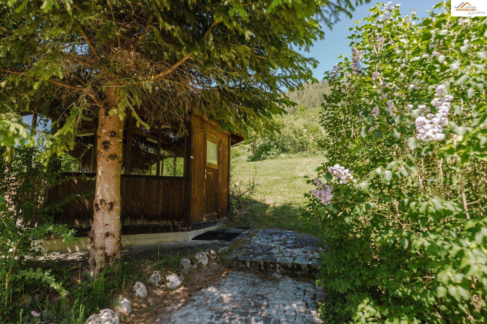 Rustikaler Holzschuppen oder Gartenhaus, umgeben von Bäumen und Büschen, mit einem Steinweg davor.