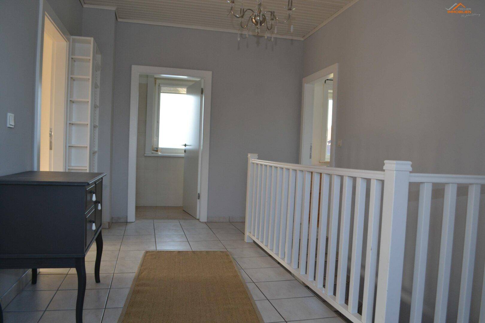 Heller Flur mit Fliesenboden, weißem Treppengeländer und einem Kronleuchter an der Decke.