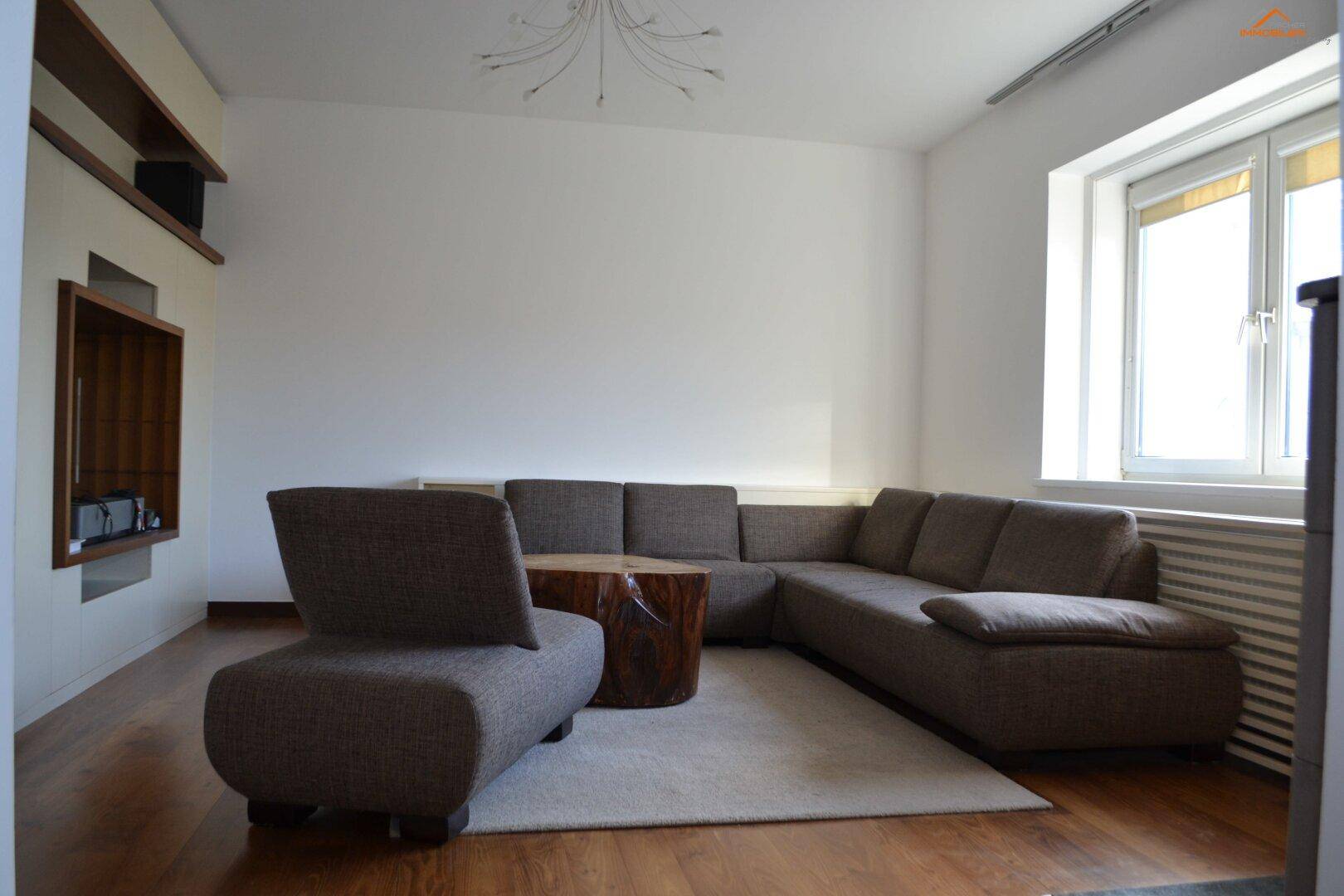 Gemütliches Wohnzimmer mit großem Ecksofa, Holzboden und viel Tageslicht durch das Fenster.