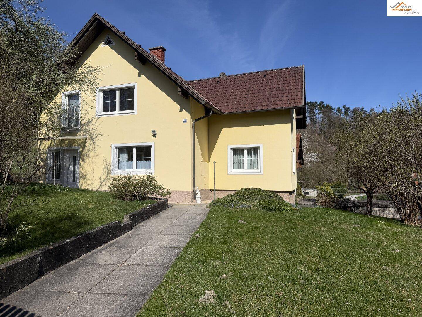 Sonniges Einfamilienhaus mit gelber Fassade und weißem Balkon vor grüner Hügellandschaft.