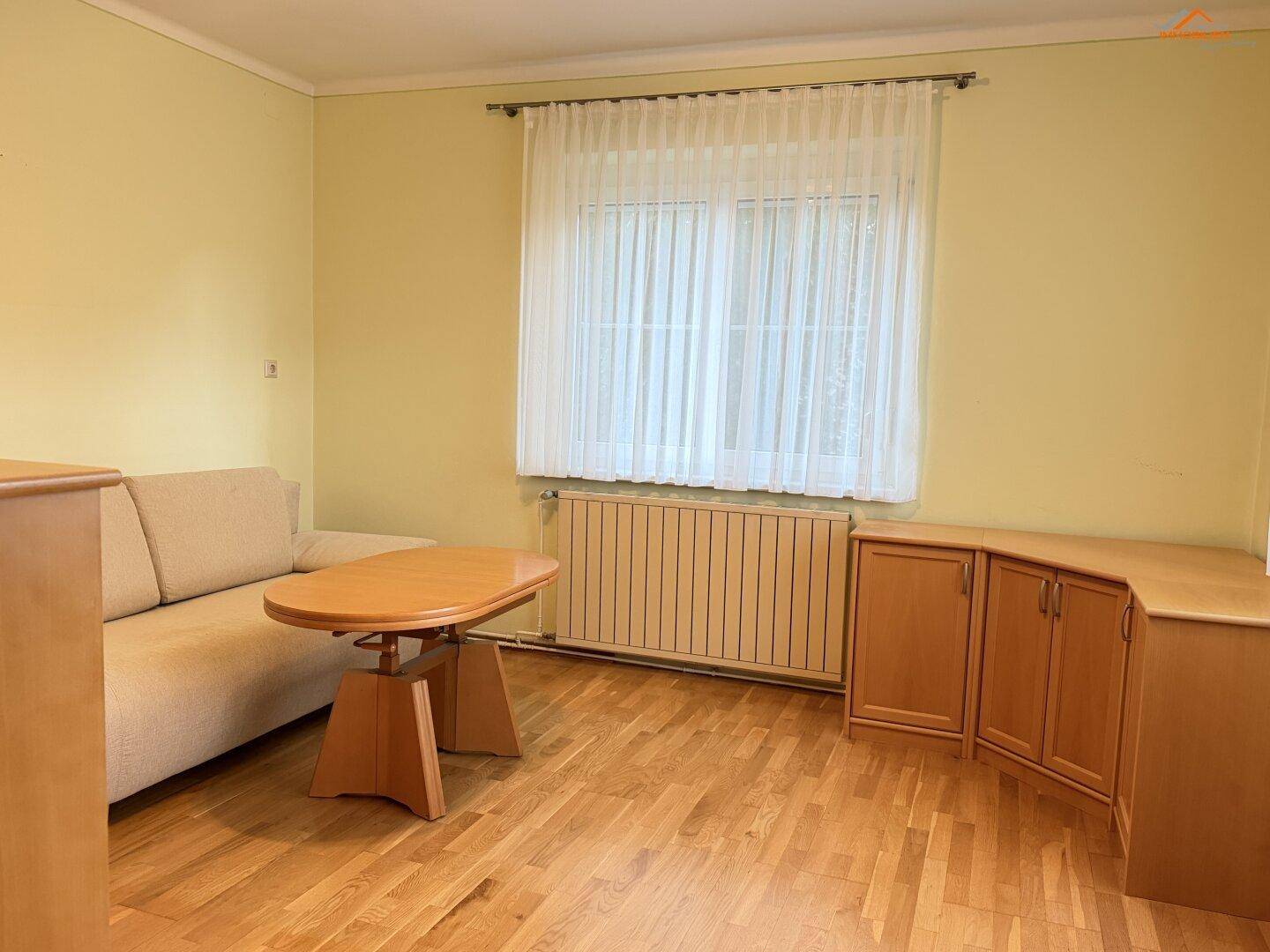 Helles Wohnzimmer mit Holzboden, Sofa und großem Fenster mit Vorhängen.