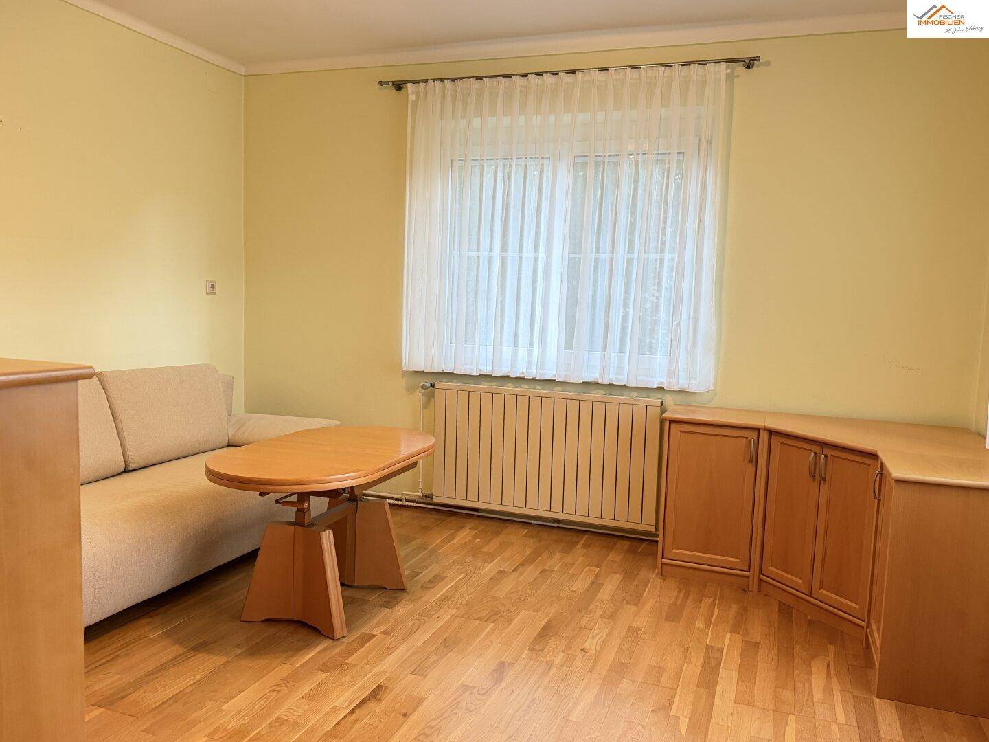 Helles Wohnzimmer mit Laminatboden, beigem Sofa und Holzmöbeln unter einem Fenster.