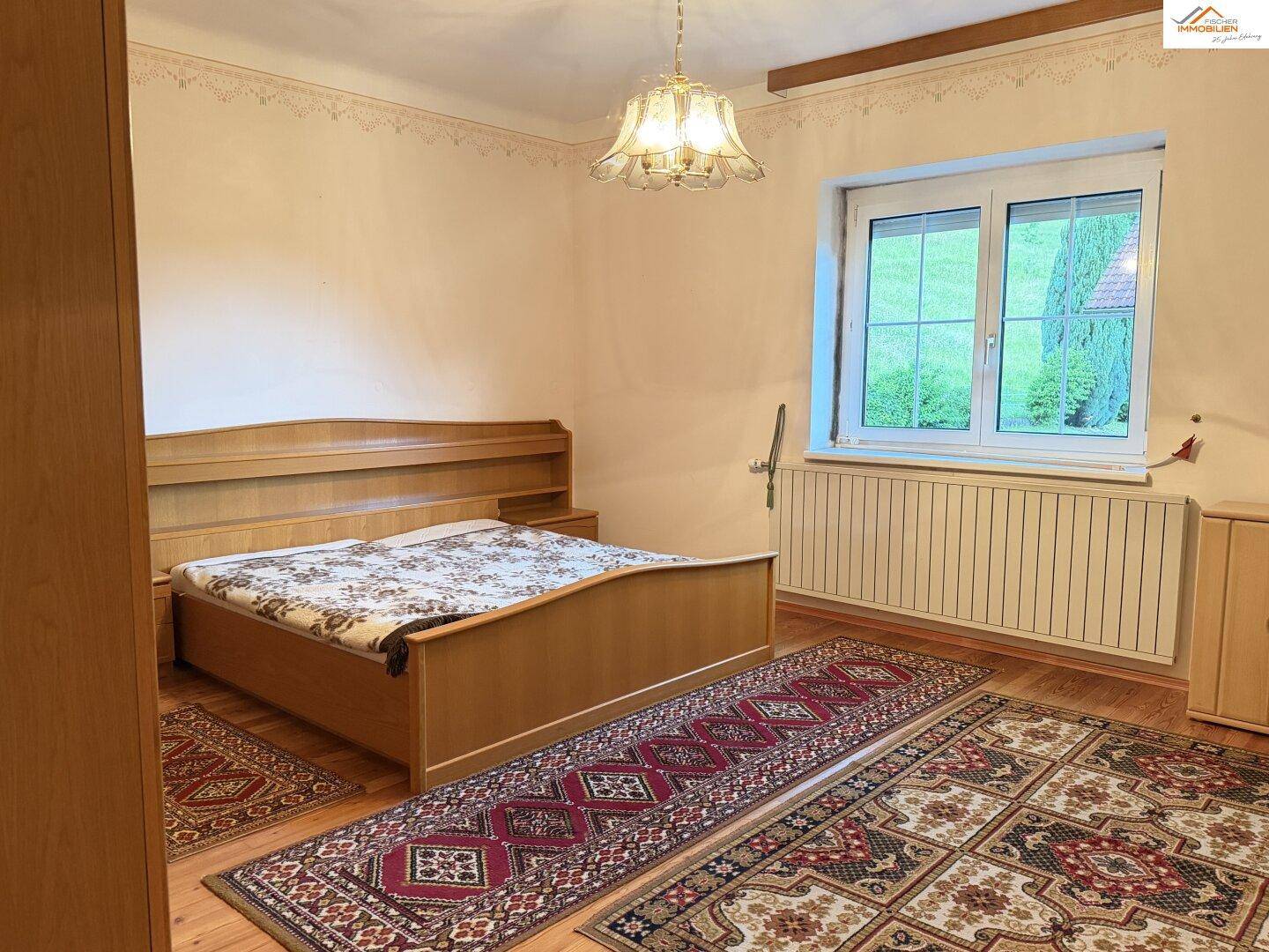 Schlafzimmer mit Holzbett, Teppichen und Blick ins Grüne durch das Fenster.