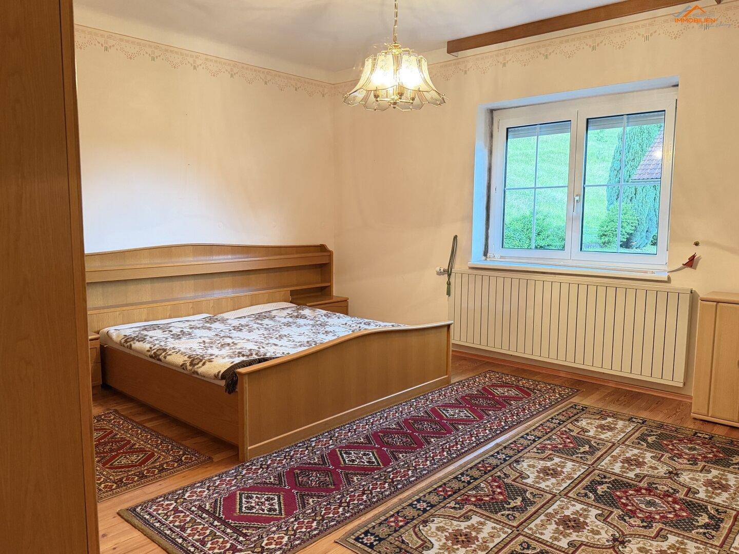 Schlafzimmer mit Holzbett, Teppichen und Fenster mit Blick ins Grüne.