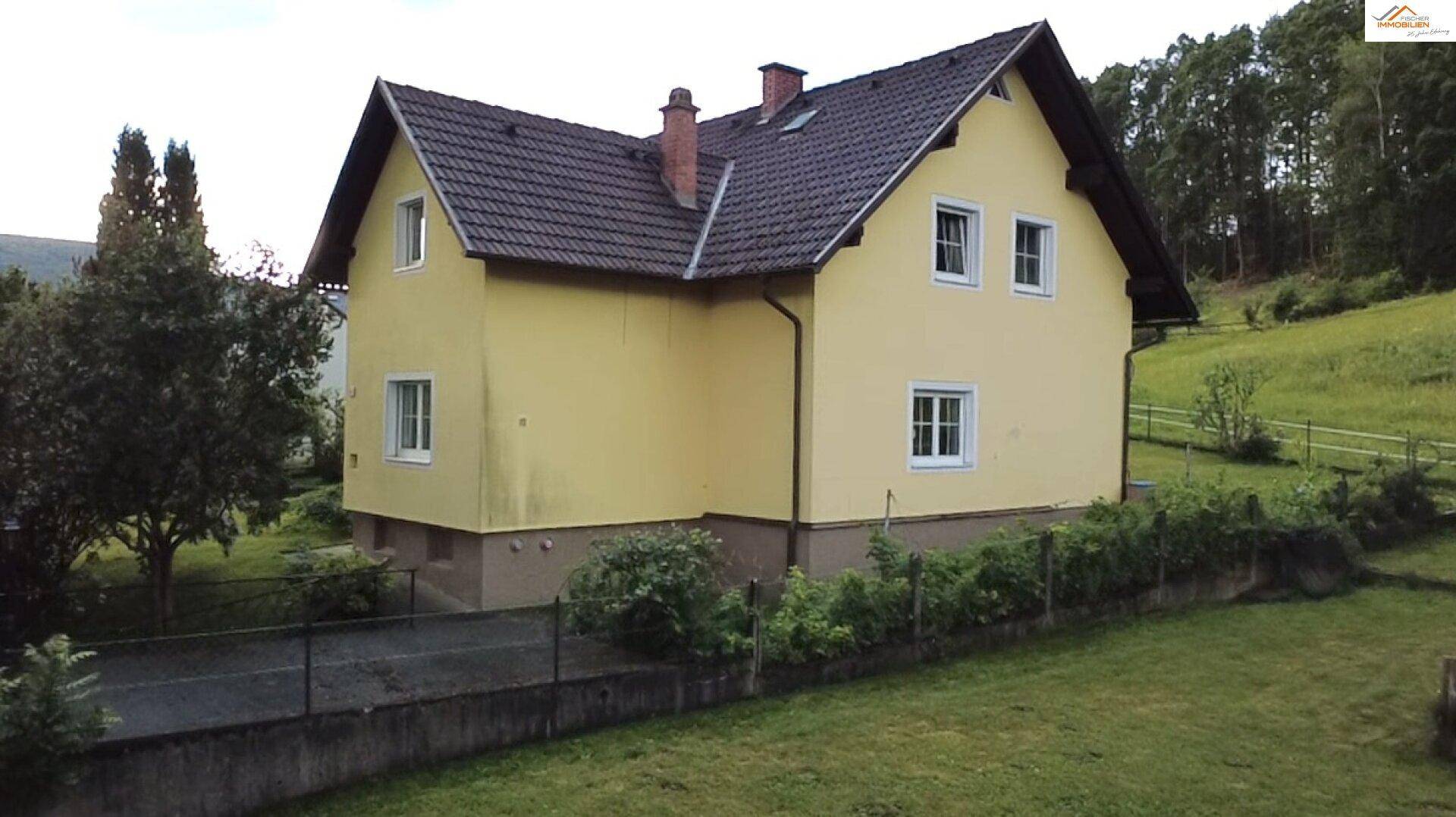 Seitenansicht des gelben Hauses mit braunem Dach und umgebender grüner Landschaft.
