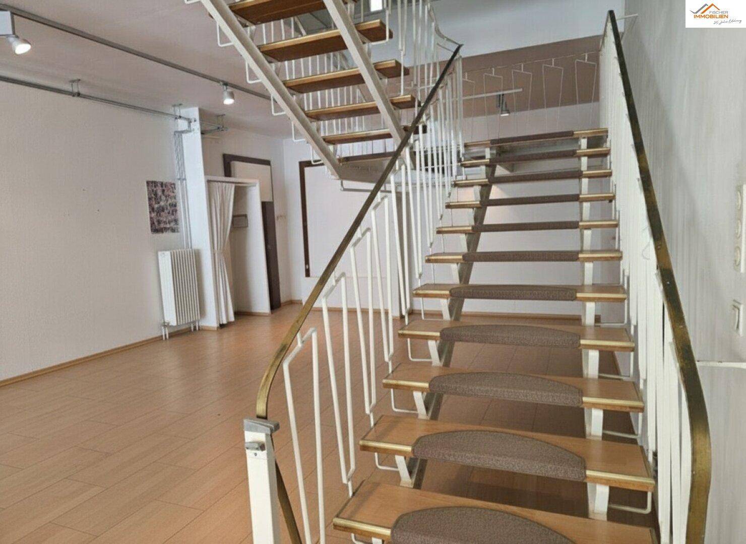 Eine geschwungene Treppe mit Teppichstufen und Metallgeländer führt in das Obergeschoss.