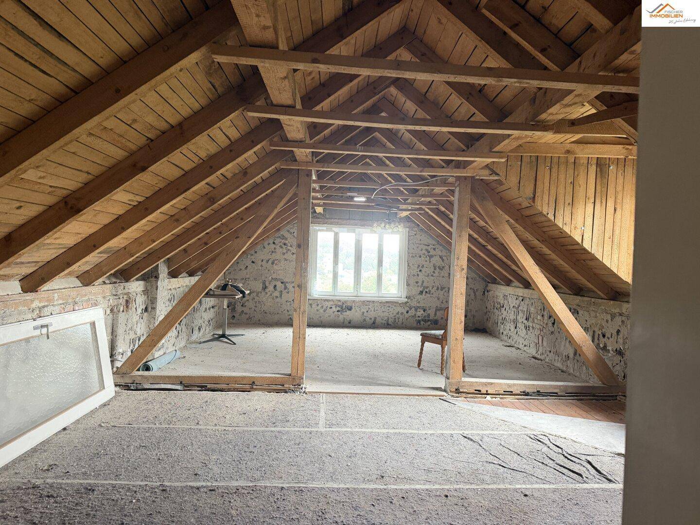 Unfertiger Dachboden mit sichtbaren Holzbalken und einem Fenster, bereit für den Ausbau.