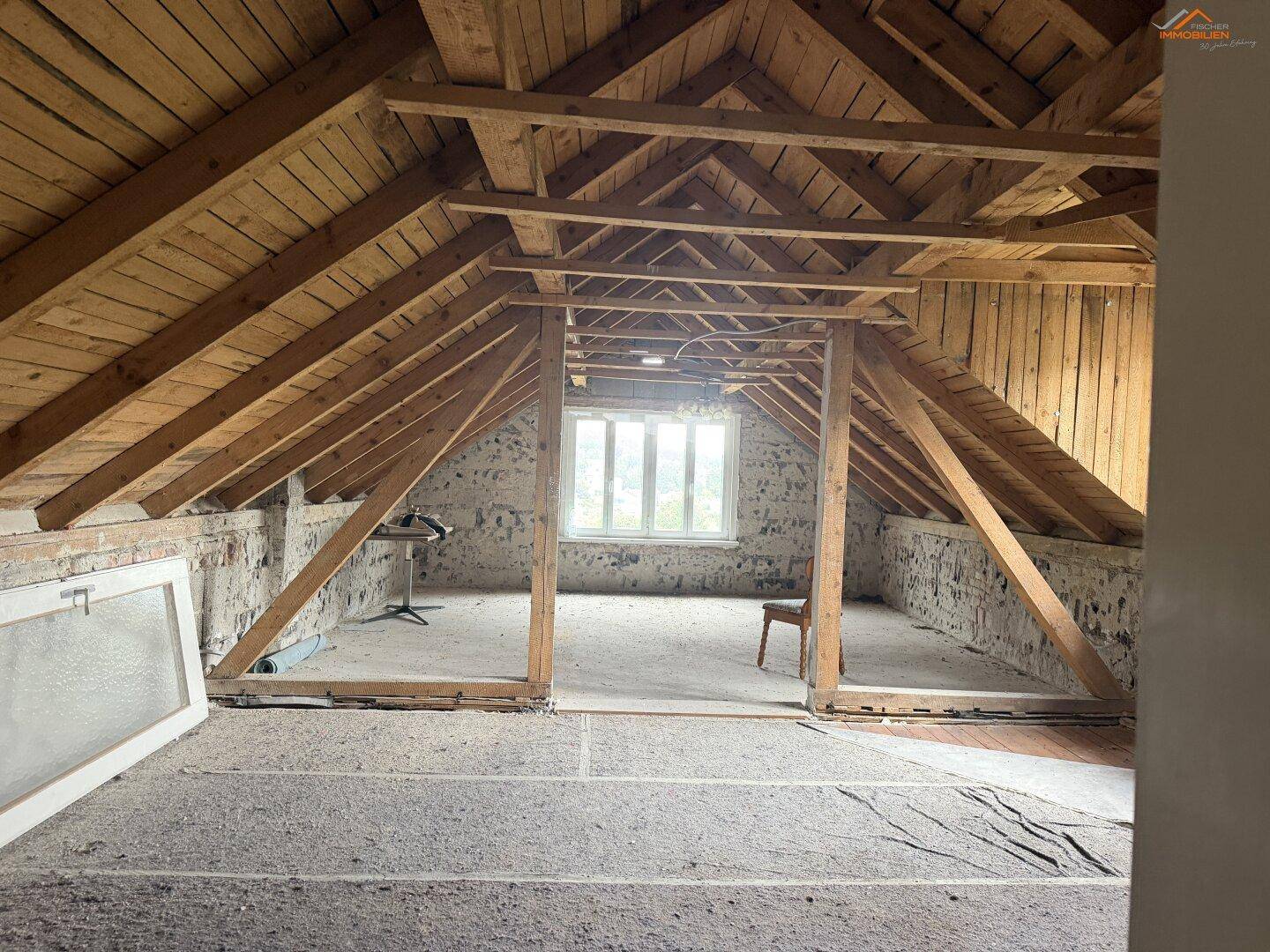 Dachbodenraum im Rohzustand mit sichtbaren Holzbalken und einem Fenster.