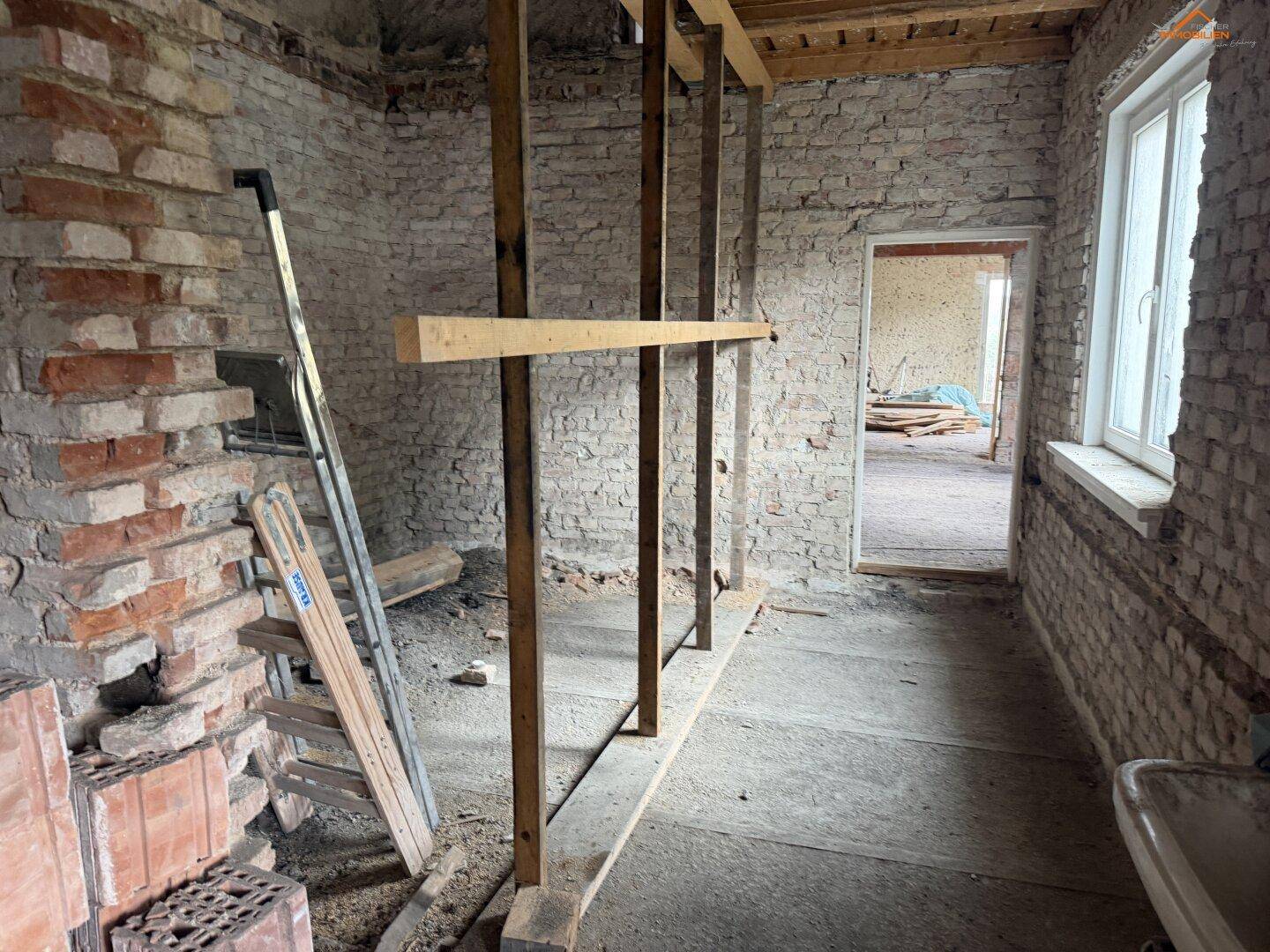 Flur im Rohbau mit unverputzten Ziegelwänden, Holzstützen und einem Fenster.