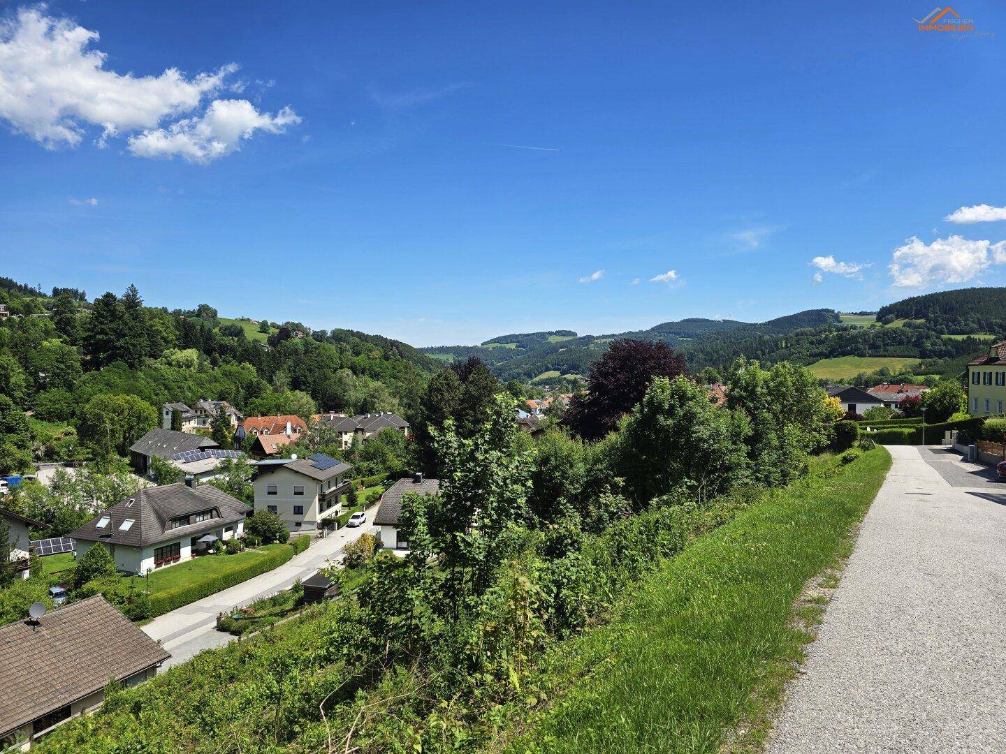 Weitläufiger Blick auf eine Wohnsiedlung in einem grünen Tal mit einer Straße und bewaldeten Hügeln.