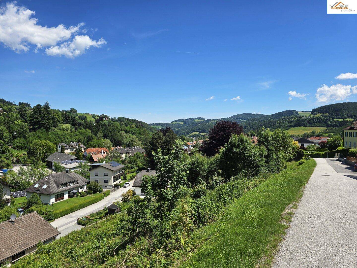 Panoramablick über eine grüne Wohnsiedlung in einer hügeligen Landschaft unter blauem Himmel.