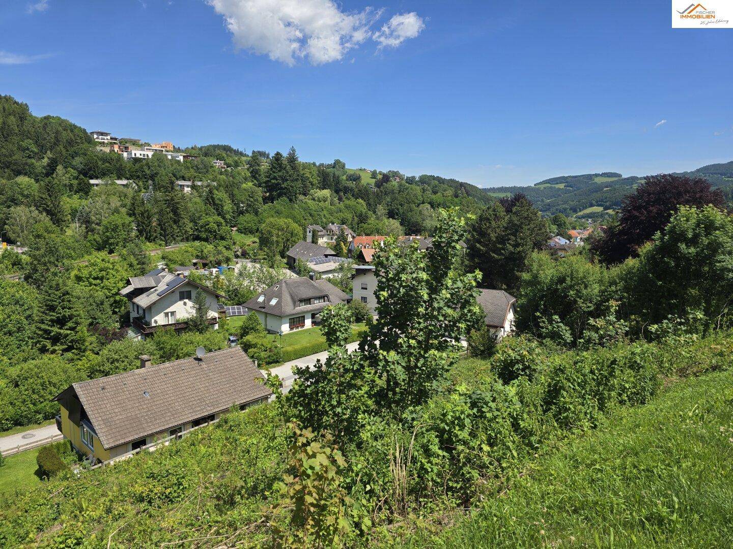 Panoramablick über eine grüne Wohnsiedlung in einer hügeligen Landschaft unter blauem Himmel.