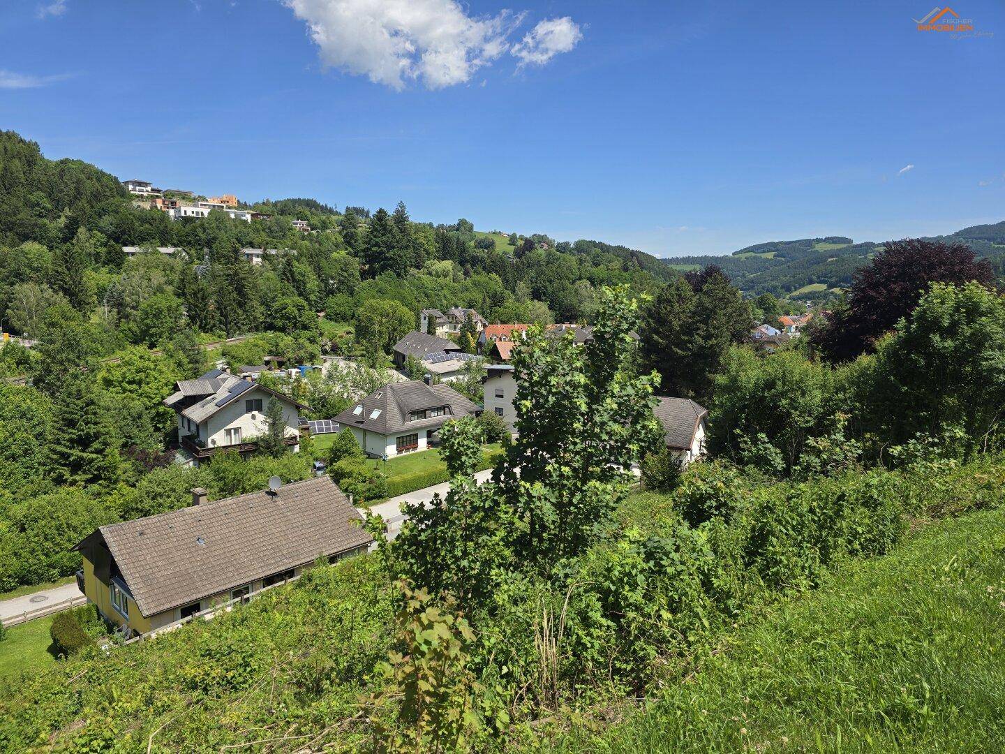 Ausblick auf eine Wohnsiedlung in einem grünen Tal, umgeben von bewaldeten Hügeln unter blauem Himmel.