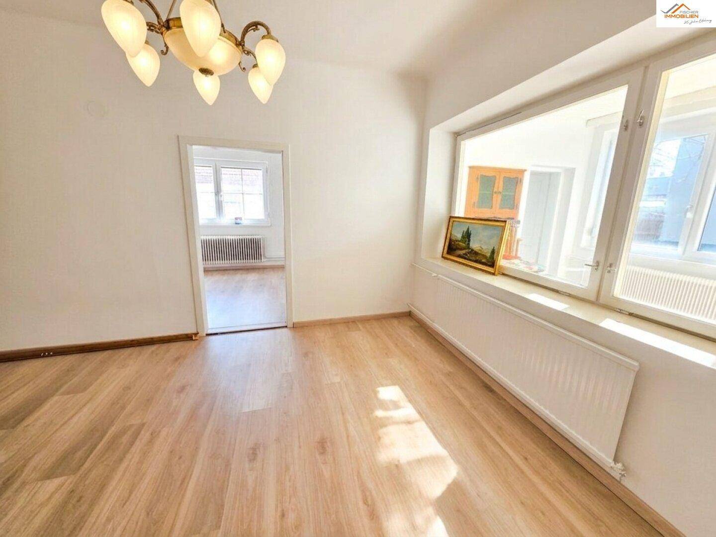 Helles Zimmer mit großen Fenstern, Holzboden und einem Kronleuchter, bereit zur individuellen Gestaltung.