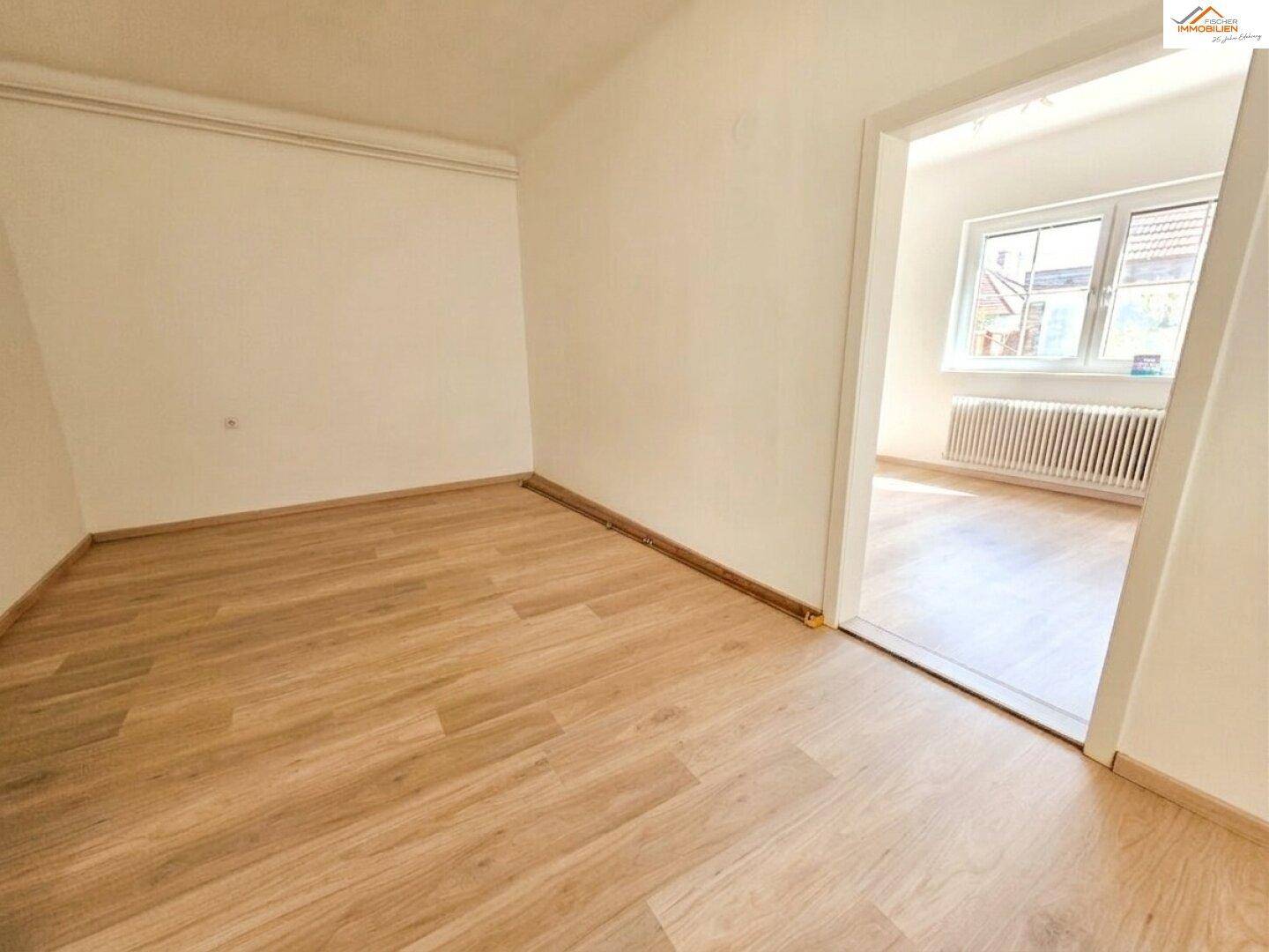 Leeres Zimmer mit hellem Holzboden und einem Durchgang zu einem weiteren Raum mit Fenster.