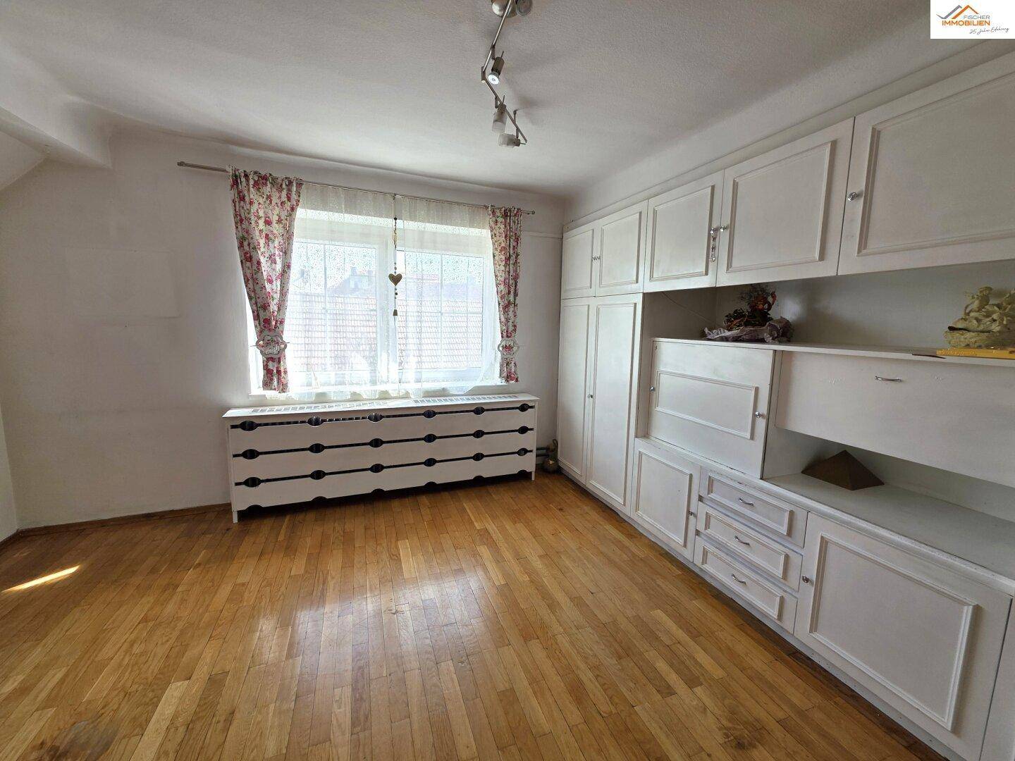Geräumiges Zimmer mit umfangreichen weißen Einbauschränken und einem Fenster mit Vorhängen.