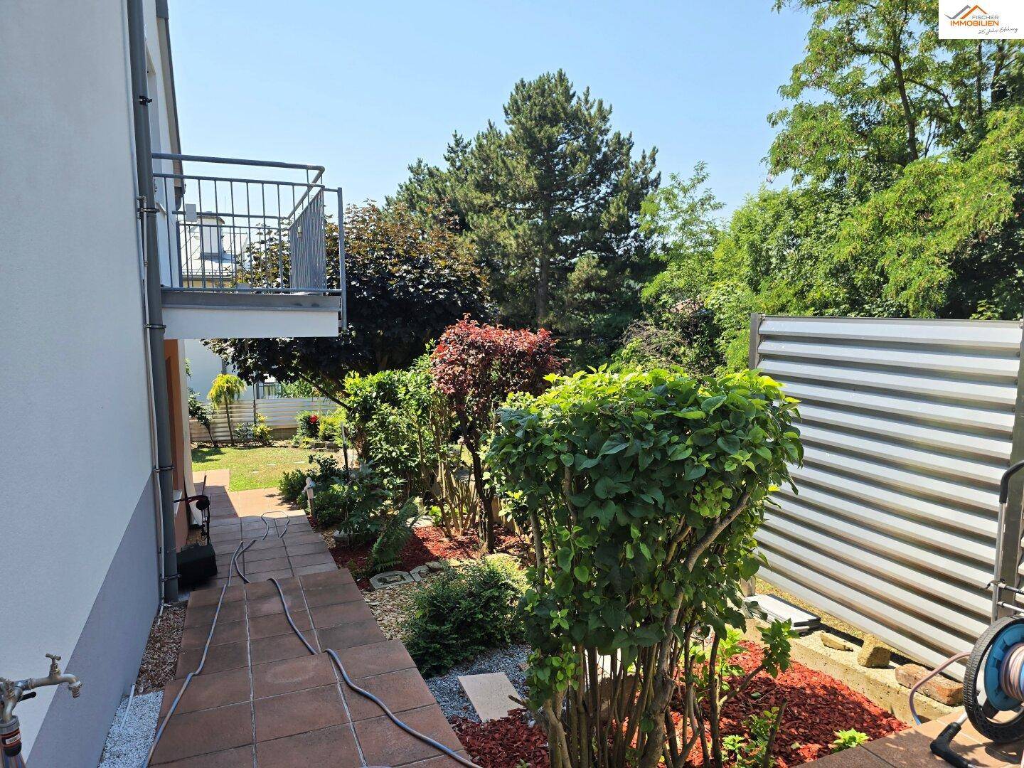 Seitlicher Gartenweg mit Bepflanzung und kleinem Balkon im Hintergrund, umgeben von Grün.