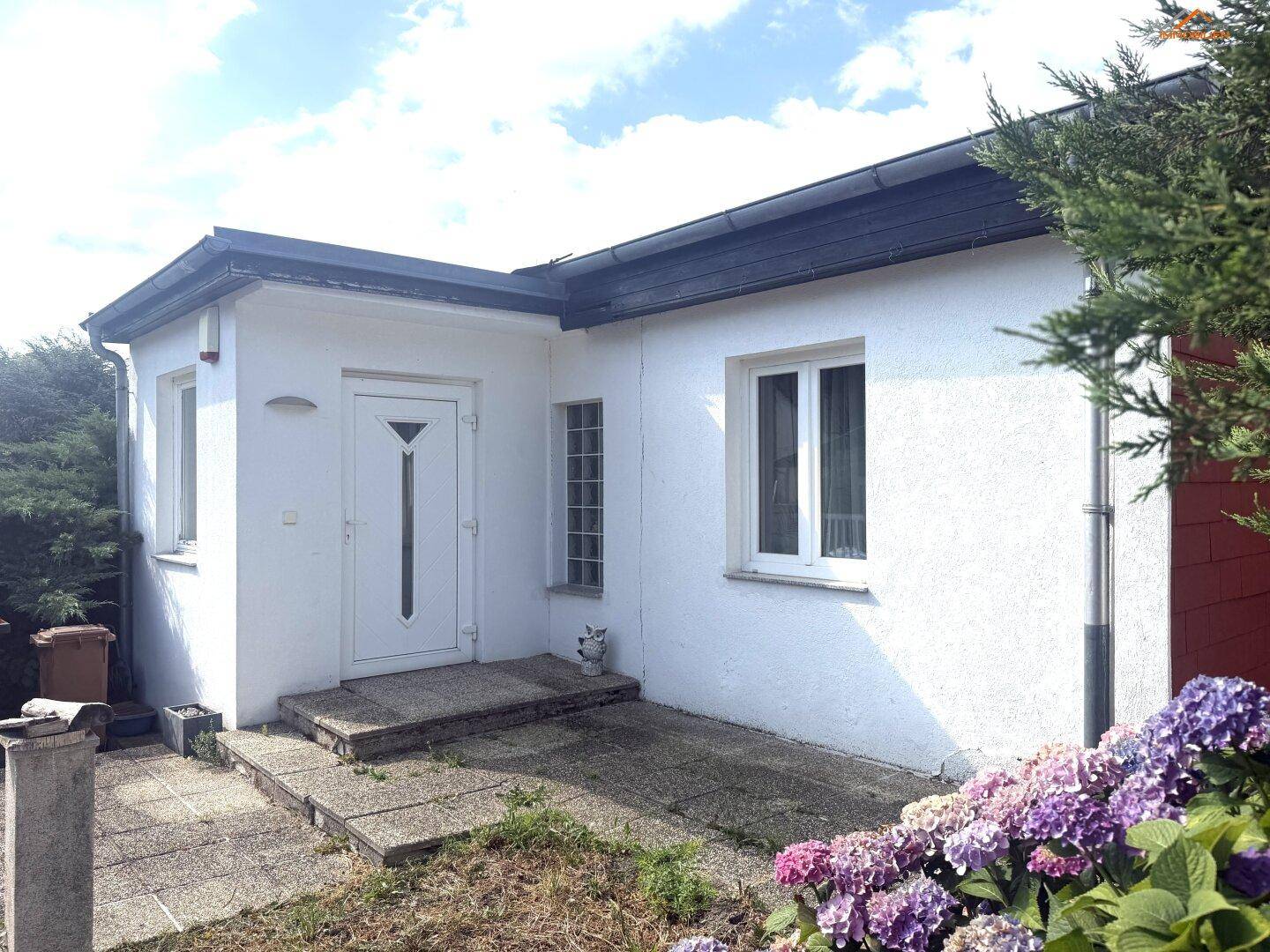 Eingangsbereich eines weißen Hauses mit Blumenbeeten und gepflastertem Zugang.