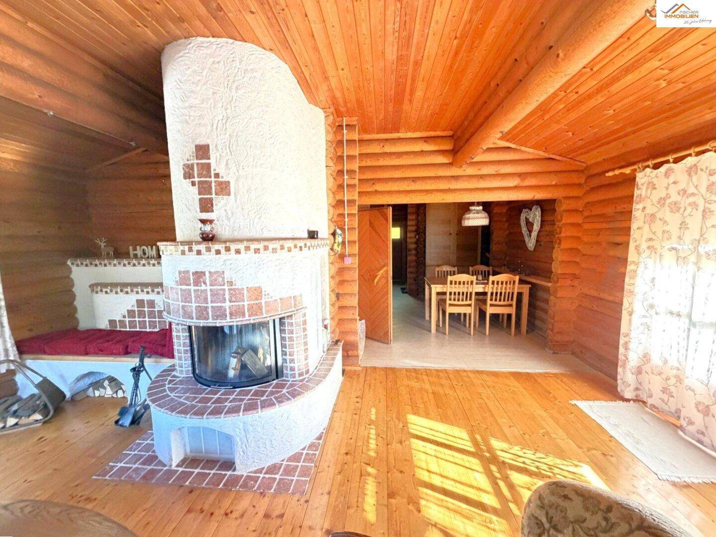 Gemütliches Wohnzimmer im Blockhaus mit großem Kamin, Holzboden und Blick auf den Essbereich.