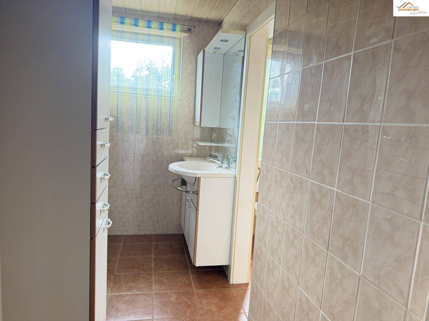 Badezimmer mit Waschbecken, Spiegelschrank und hellbraunen Fliesen an Wänden und Boden.