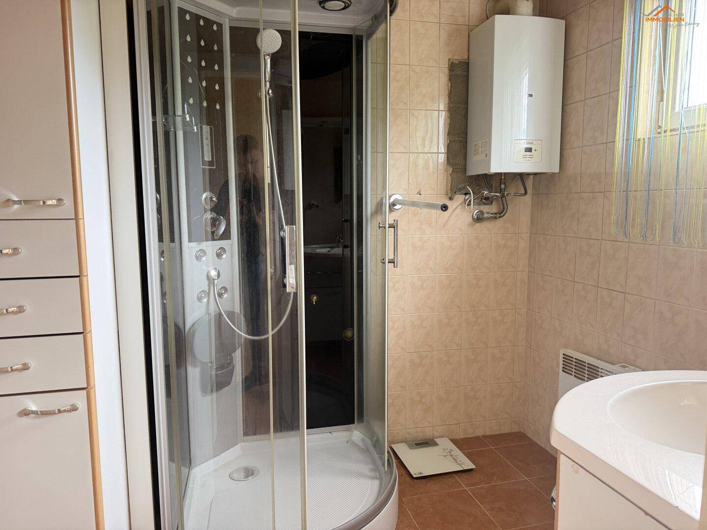 Badezimmer mit Duschkabine, Warmwasserbereiter und beigefarbenen Fliesen.