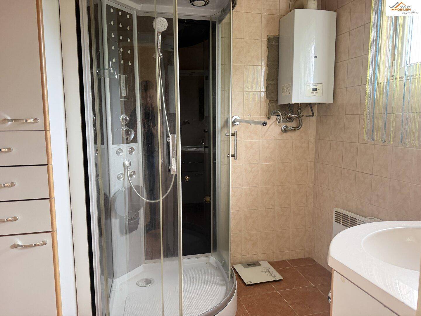 Badezimmer mit Duschkabine, Warmwasserbereiter und hellbraunen Wandfliesen.