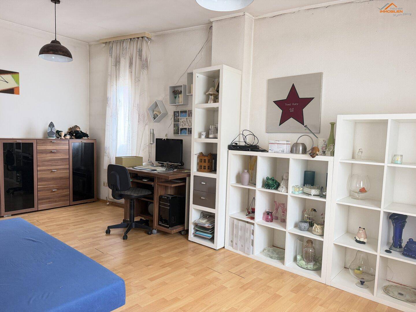Geräumiges Zimmer mit Schreibtisch, Regalen und hellem Laminatboden.