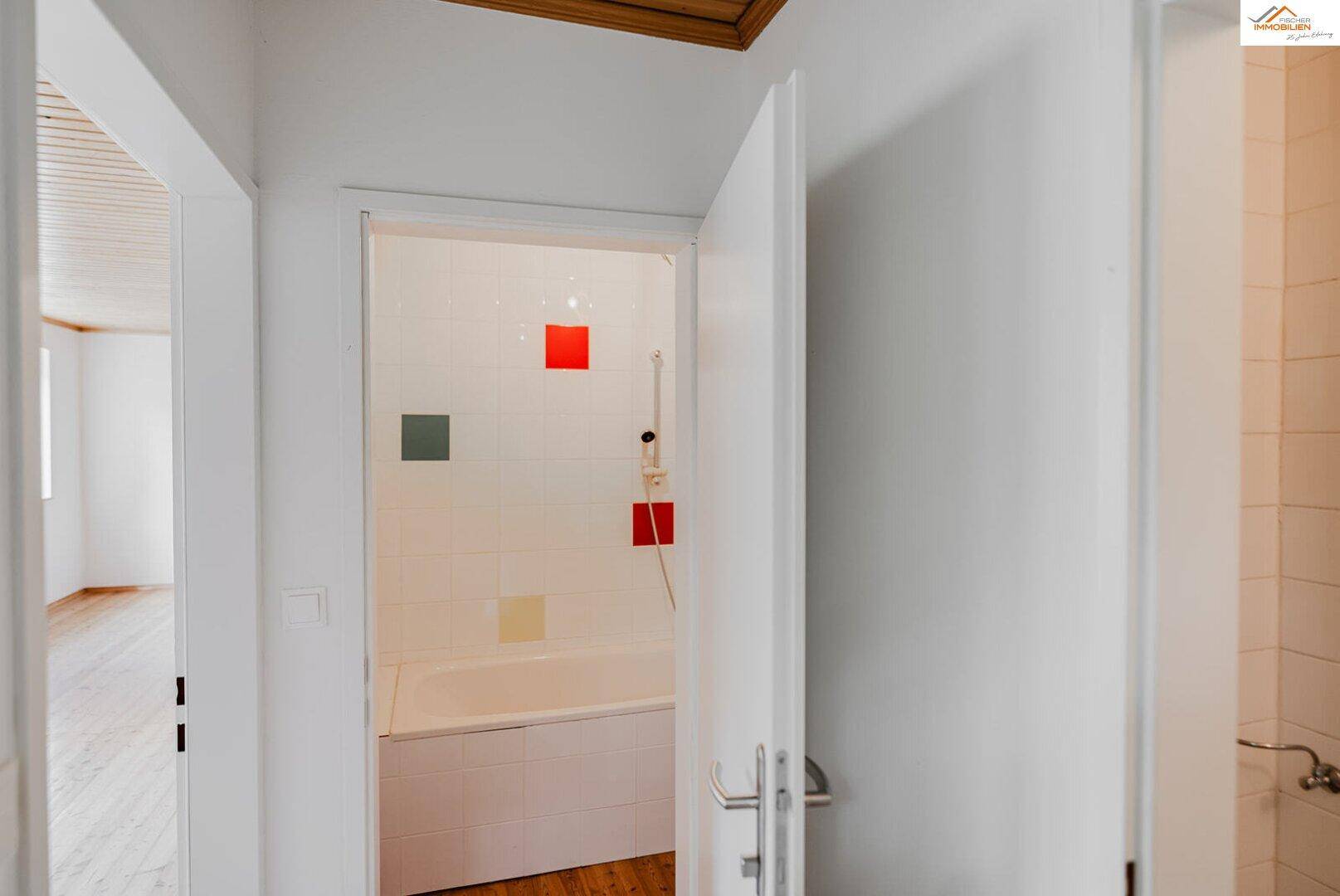 Badezimmer mit Badewanne und bunten Fliesenakzenten, sichtbar durch eine offene Tür.