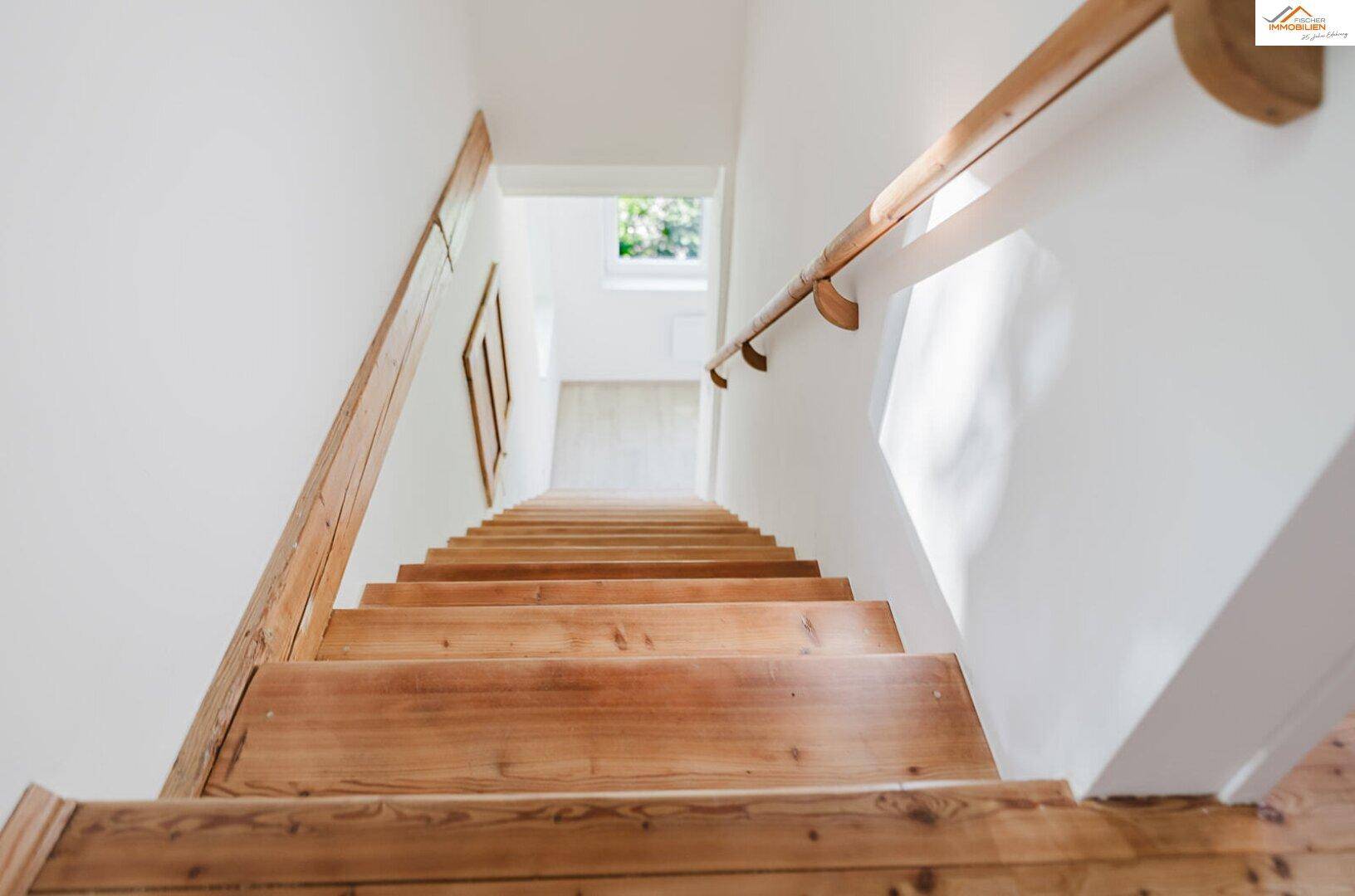 Holztreppe mit Geländer führt nach oben, beleuchtet durch ein Dachfenster.