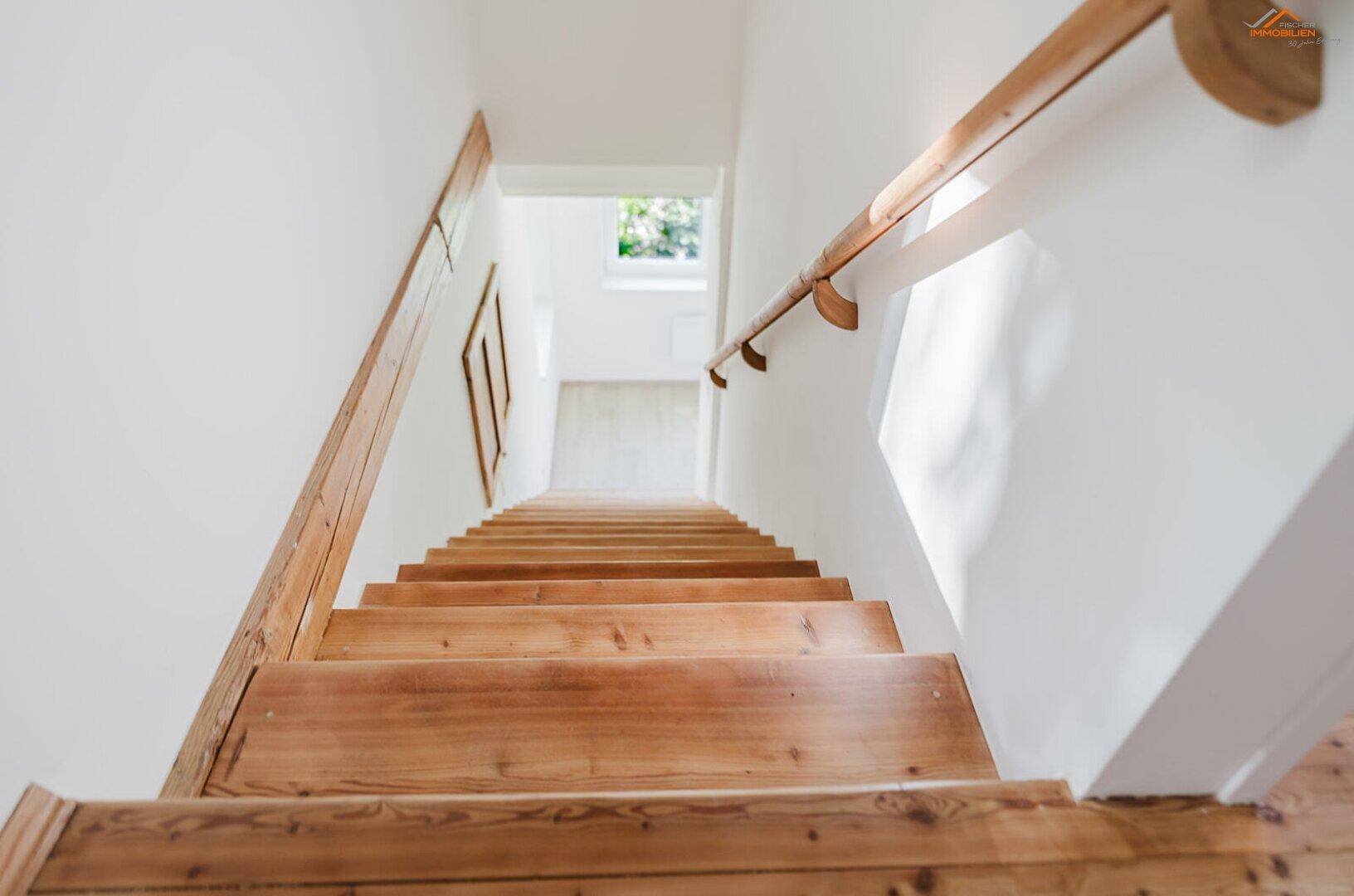 Holztreppe mit Handlauf führt nach oben, beleuchtet durch natürliches Licht.