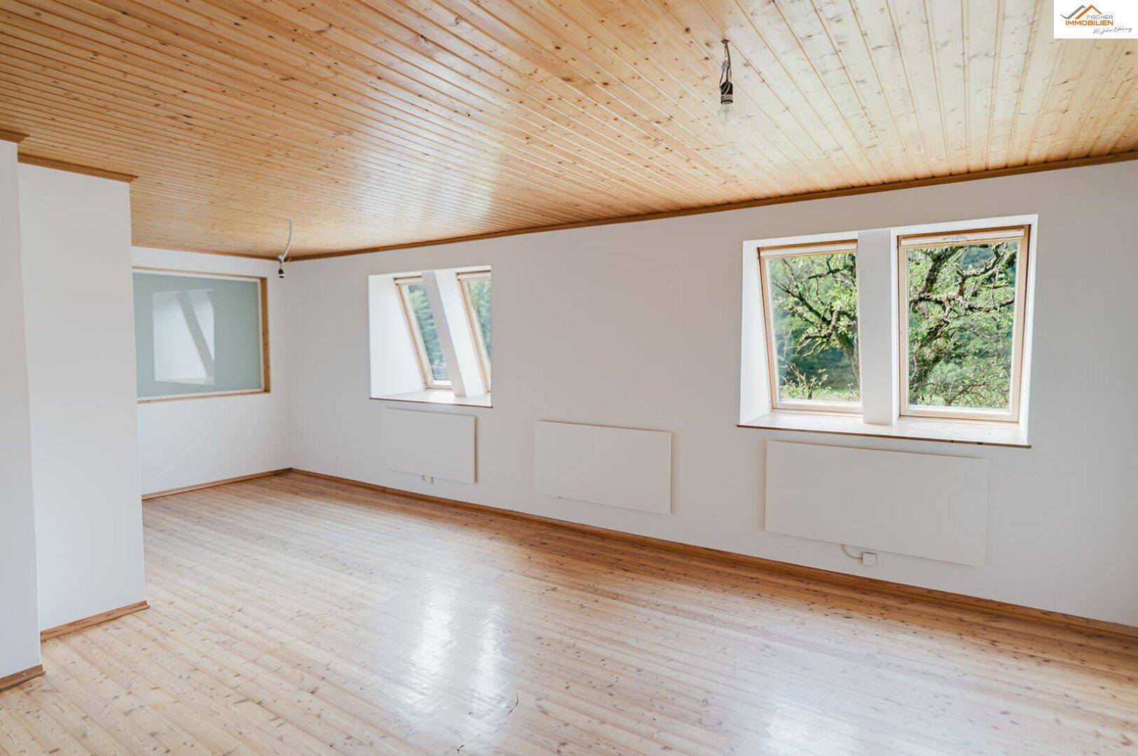 Geräumiger Raum im Dachgeschoss mit Holzdecke, Holzboden und Dachfenstern mit Waldblick.