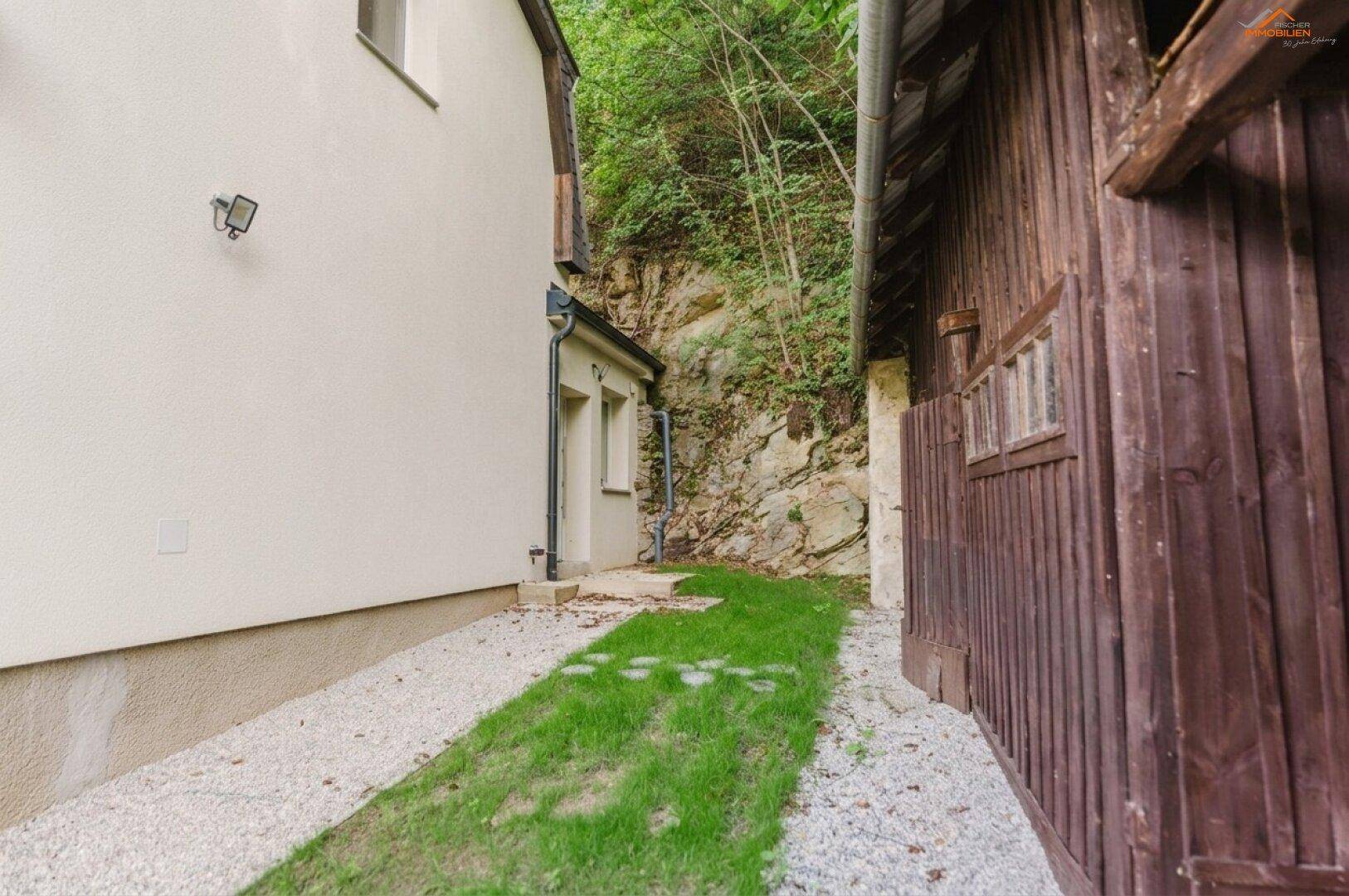 Seitenansicht des Hauses und der Holzscheune, umgeben von Kies und Grünflächen.