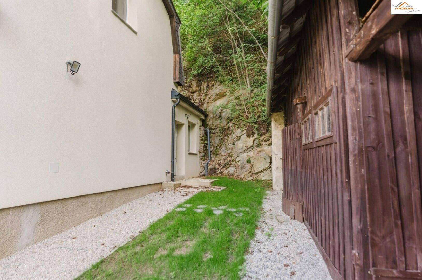 Seitenansicht des Hauses und des Holzschuppens mit einem schmalen Rasenstreifen.